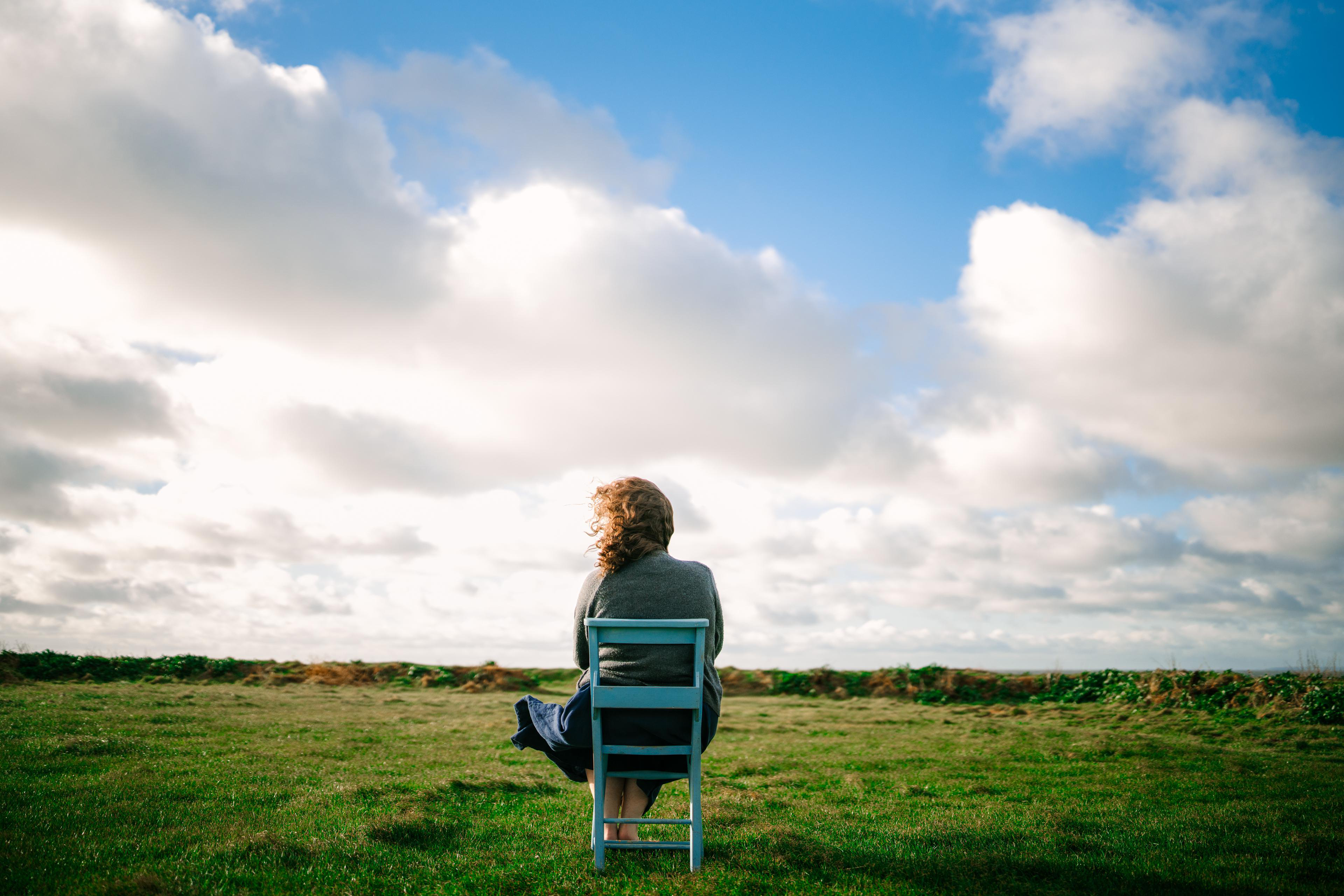 Eine Frau sitzt auf einem blauen Stuhl in einem Feld unter einem bewölkten Himmel.