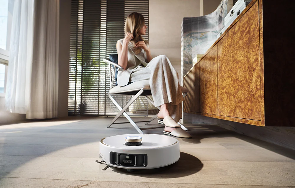 Egy fehér robotporszívó tisztít egy fapadlót egy modern szobában ülő nő előtt.