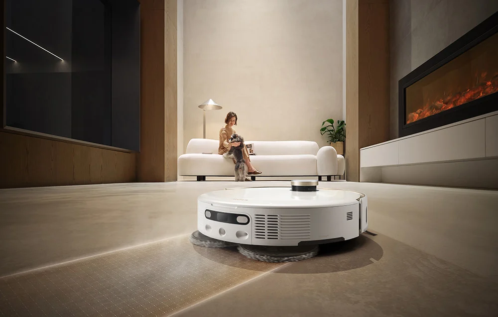 Egy fehér robotporszívó modern nappali padlóját tisztítja. Egy nő kutyával ül egy kanapén.