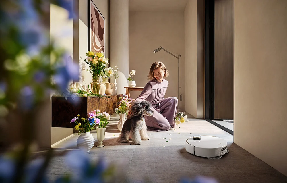 Egy lila ruhás nő egy kutya mellett térdel egy világos szobában, virágokkal és robotporszívóval.