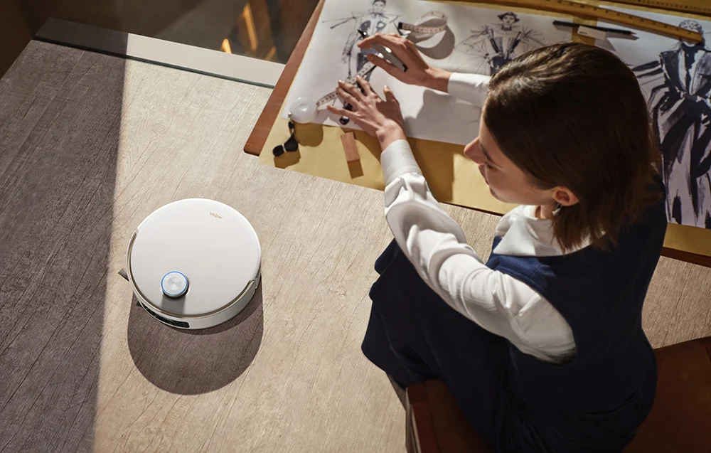 Egy fehér robotporszívó áll egy fapadlón egy divatterveken dolgozó személy mellett.