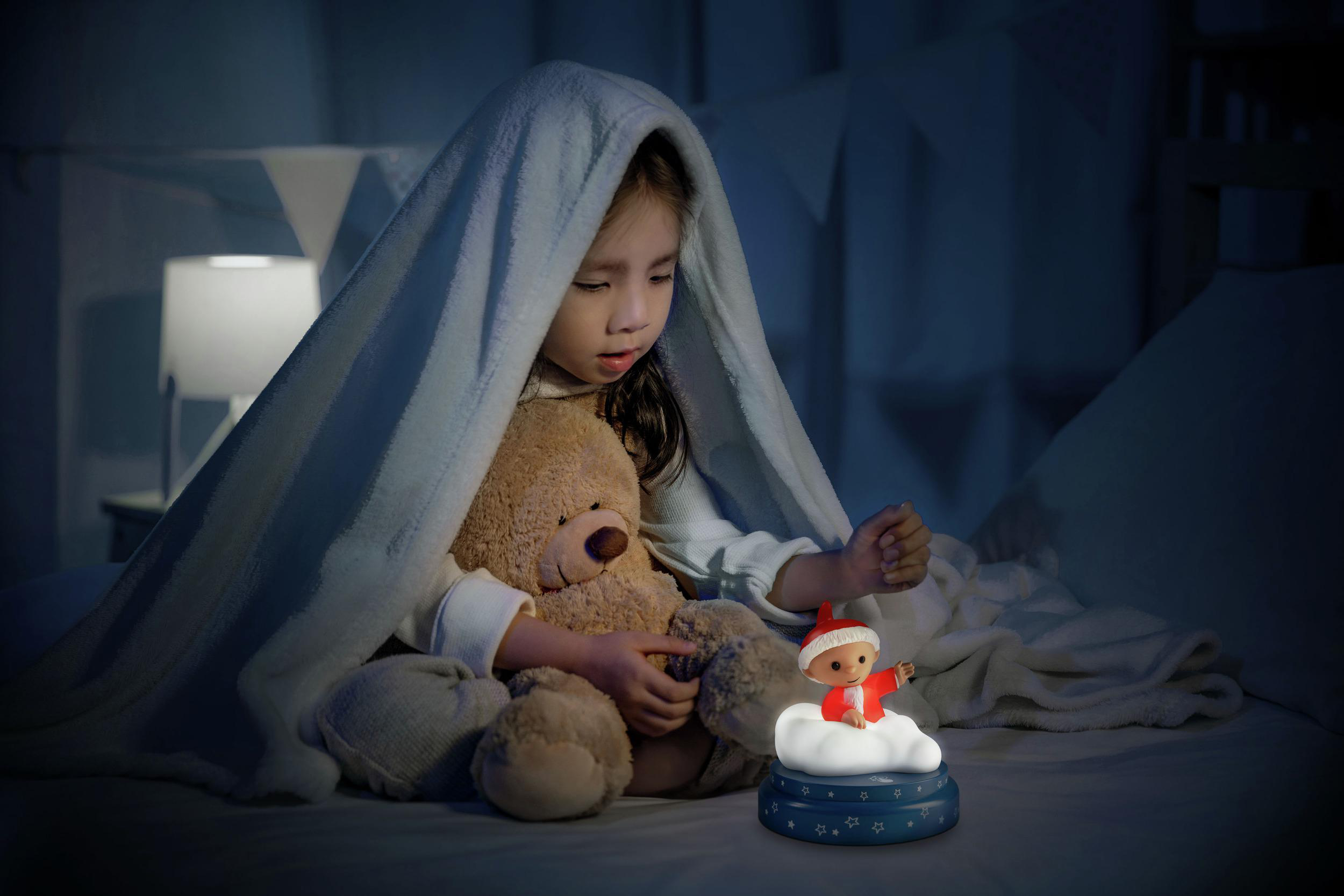 Ein kleines Mädchen mit Teddybär unter einer Decke, daneben ein Nachtlicht.