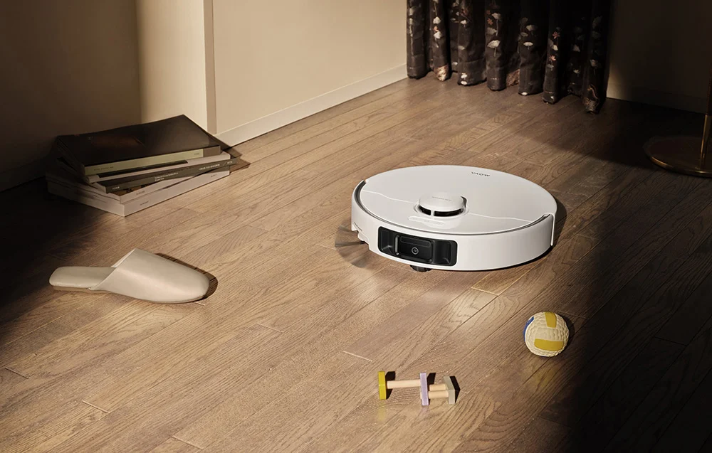 Egy fehér robotporszívó fa padlón, papucs és játékok mellett.