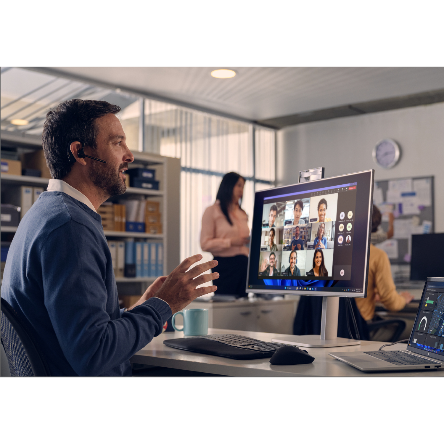 Man met headset in kantoor op videogesprek. Meerdere mensen zichtbaar op scherm.