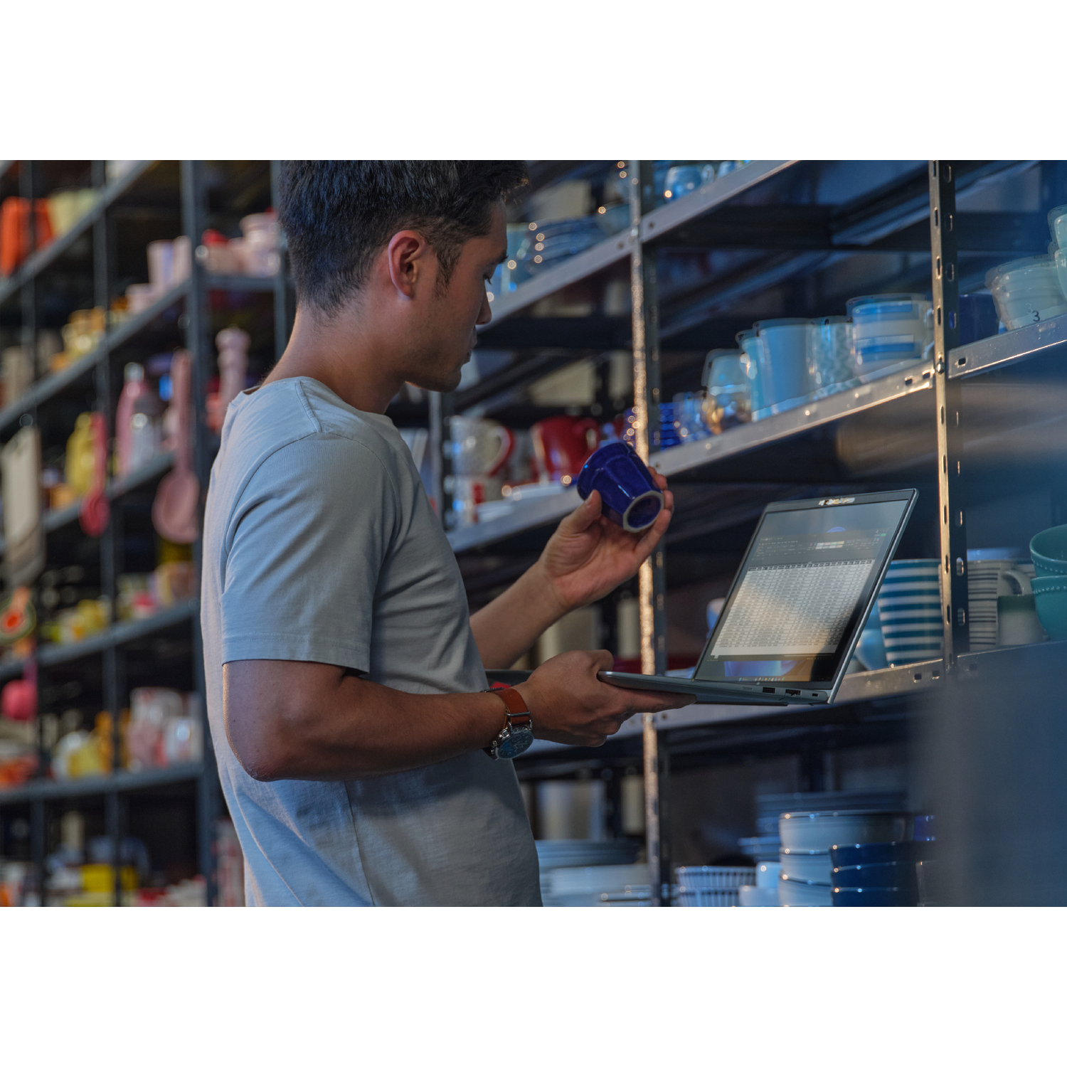 Man controleert blauwe beker met laptop in magazijnstellingen vol servies.
