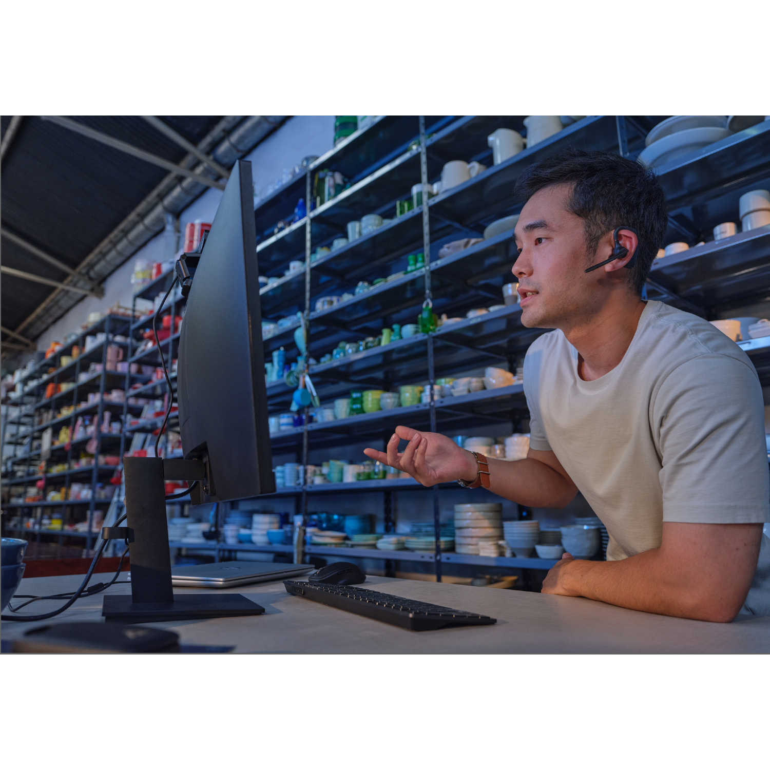 Man met headset praat bij computer in magazijn met aardewerk op rekken.