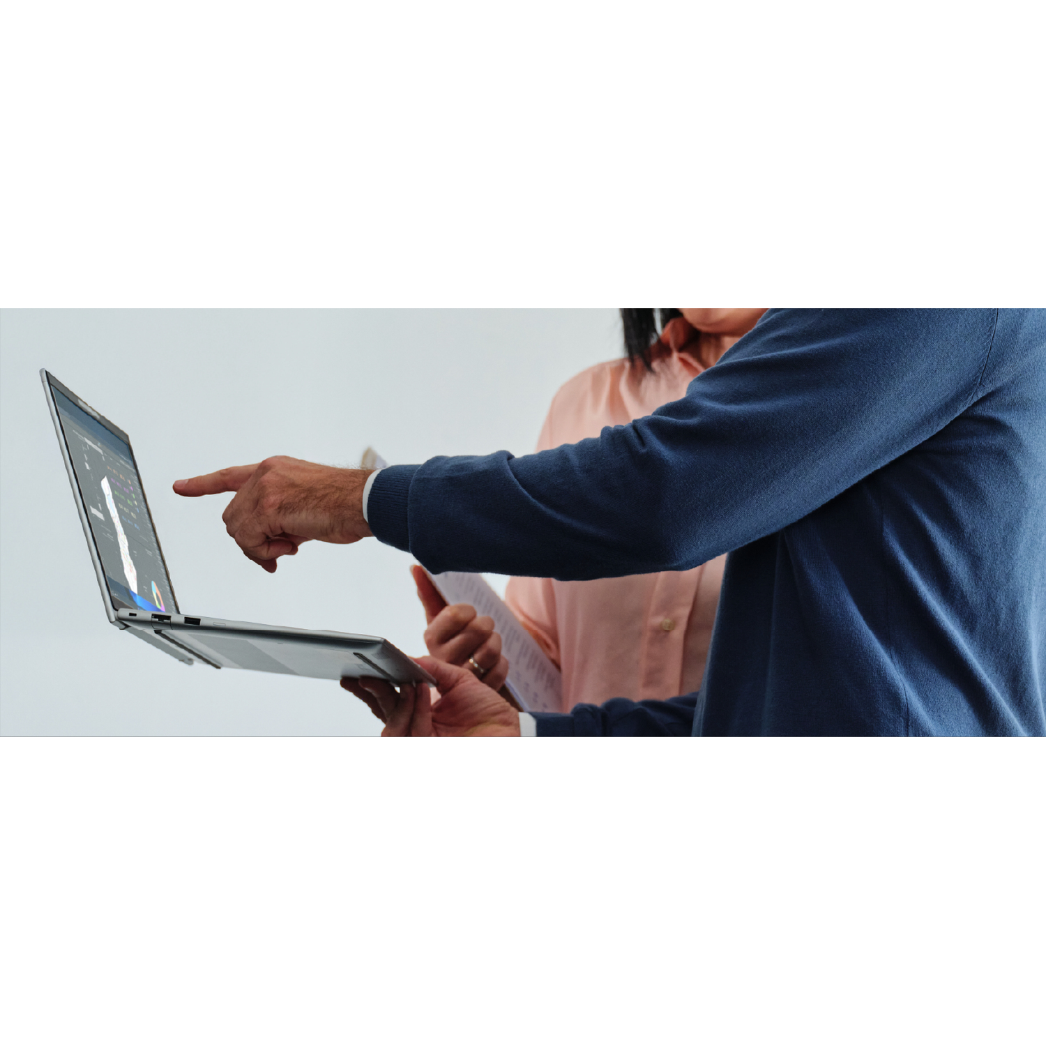 Man points at laptop, woman in pink blouse in background.