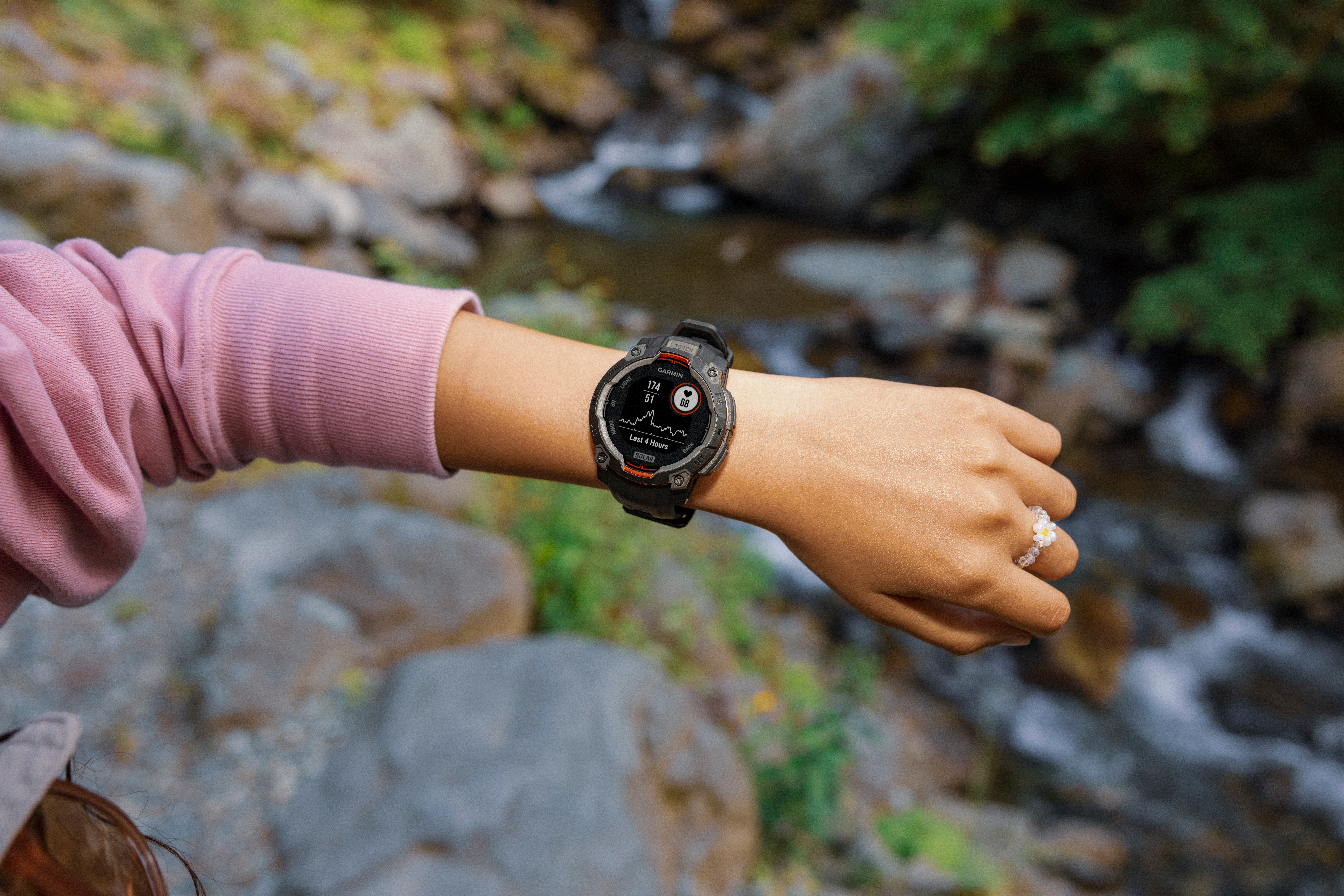 Braccio con orologio Garmin e anello fiore di perline vicino a un ruscello.
