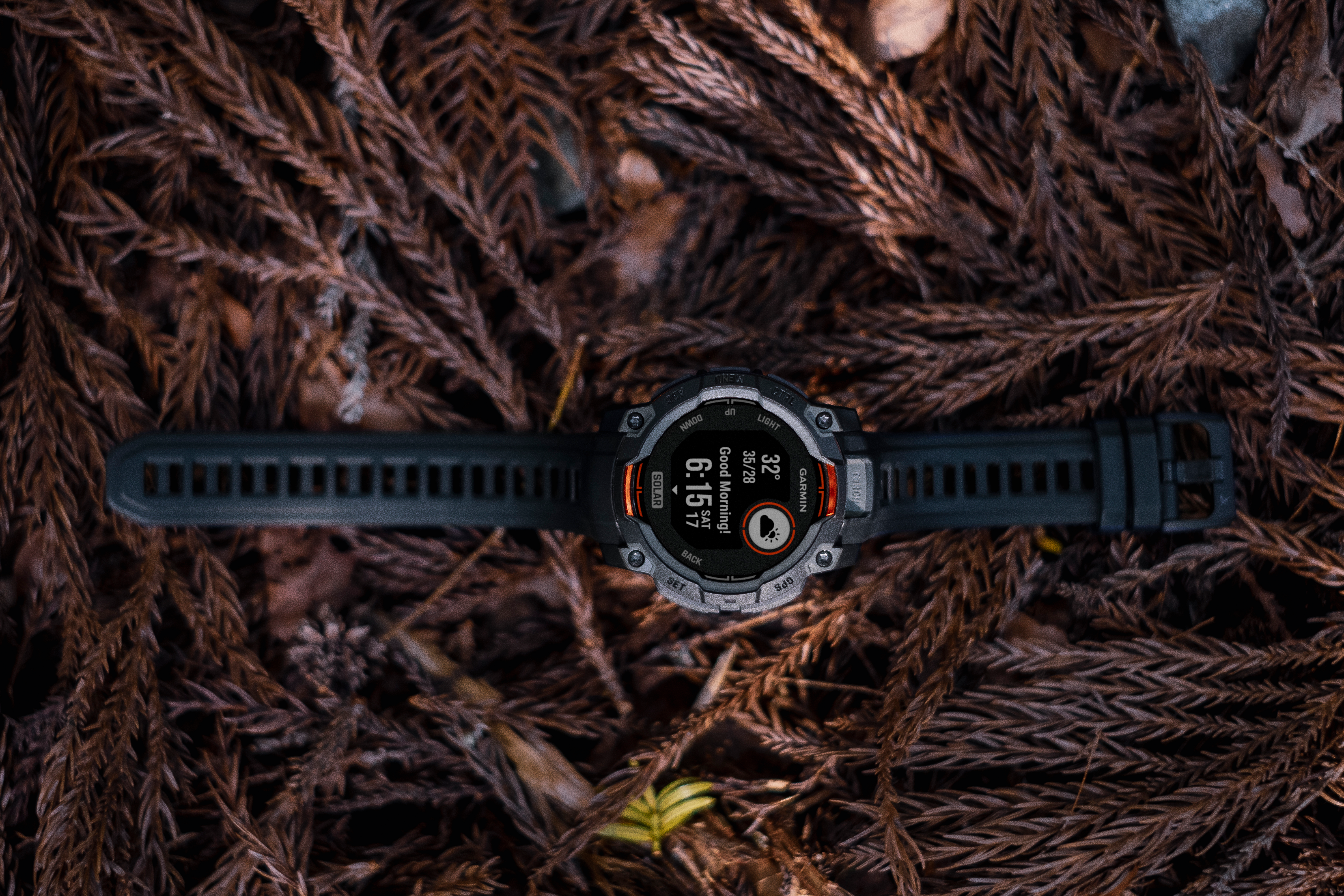 Uno smartwatch Garmin nero riposa su aghi di pino marroni. L'orologio mostra 32 gradi e le 6:15.
