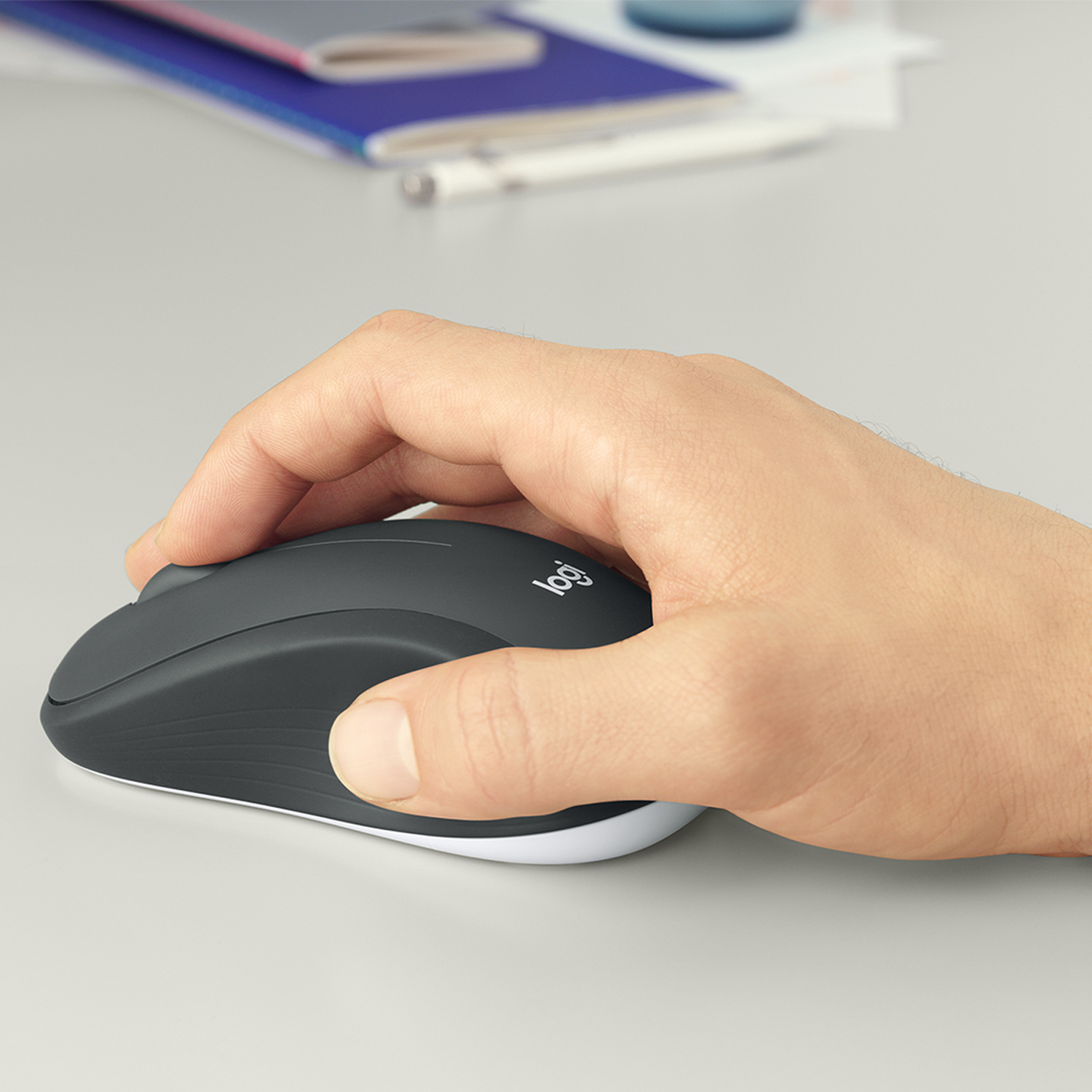 Dłoń trzyma ciemnoszarą mysz Logitech na jasnoszarym biurku. Notatniki w tle.