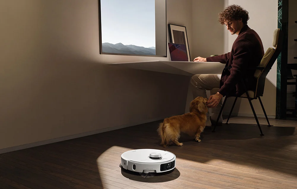 Férfi az asztalnál simogatja a kutyát. Robotporszívó tisztítja a padlót.