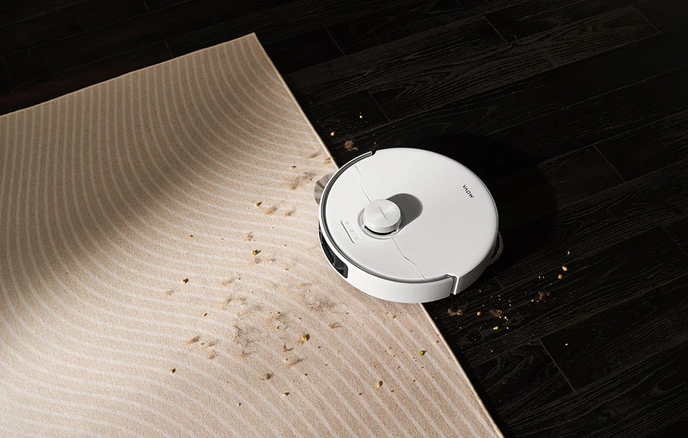 Egy fehér robotporszívó csíkos szőnyeget és sötét fapadlót tisztít, törmelékkel.