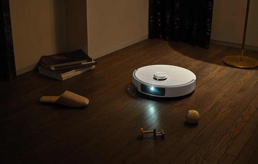Egy fehér robotporszívó tisztít egy fapadlót, mellette papucs és játékok.