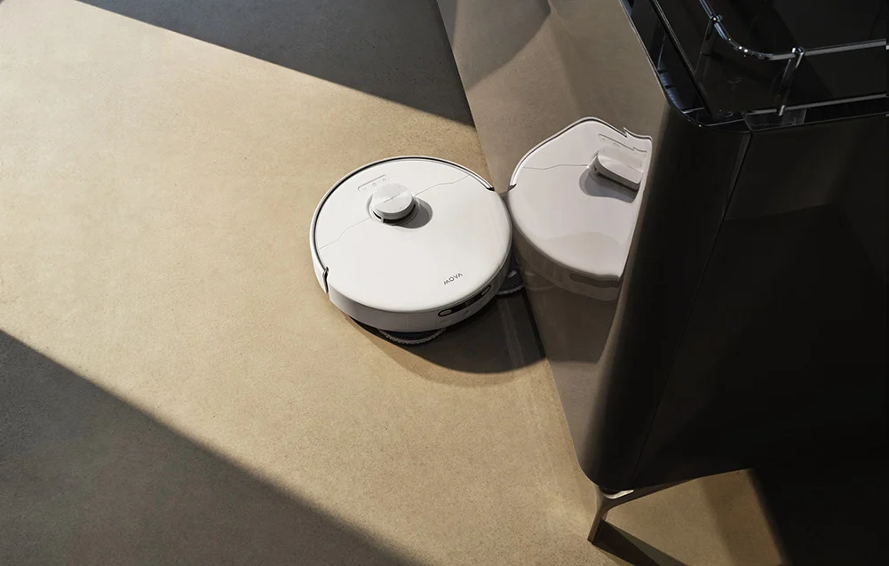 Egy fehér robotporszívó áll egy sötét egység mellett.