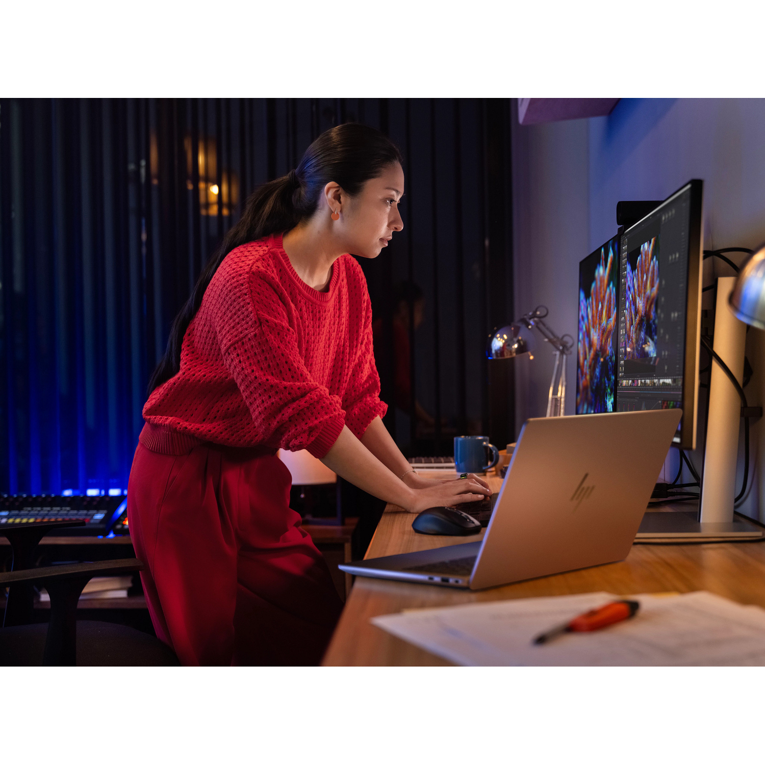 A woman in a red sweater and pants works on a laptop and computer.