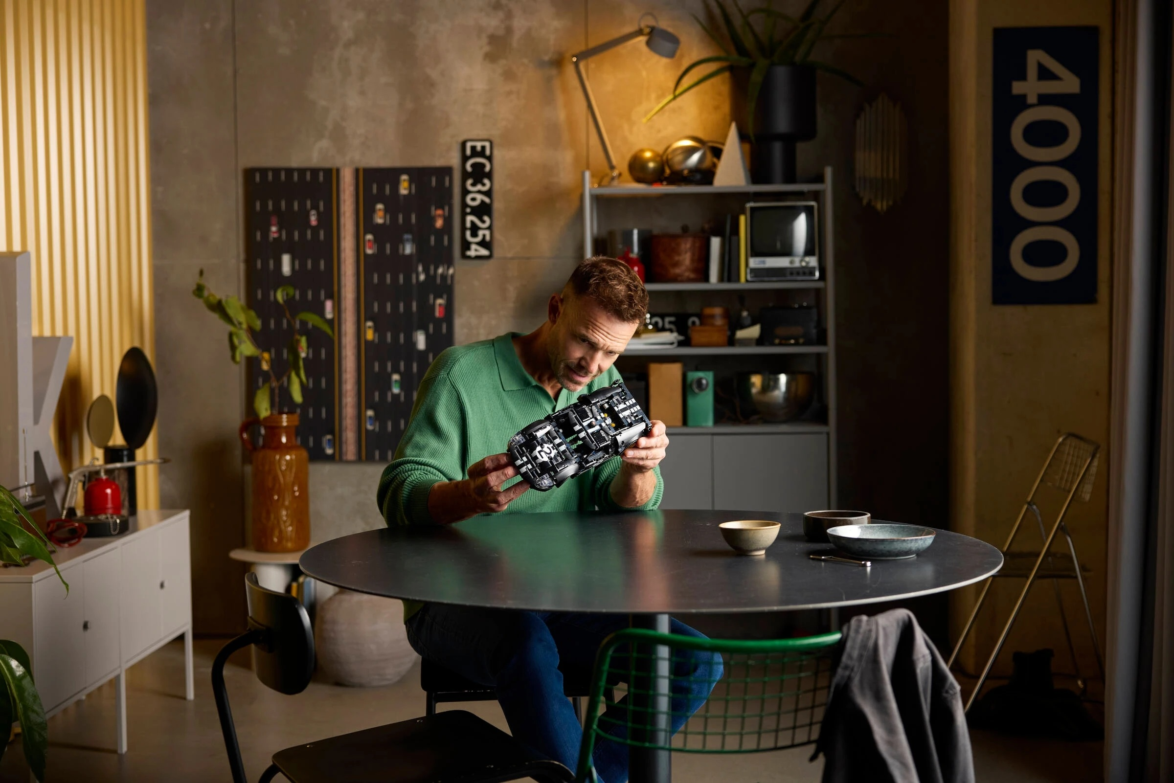 Mężczyzna w zielonym swetrze trzyma czarny model samochodu przy okrągłym stole.