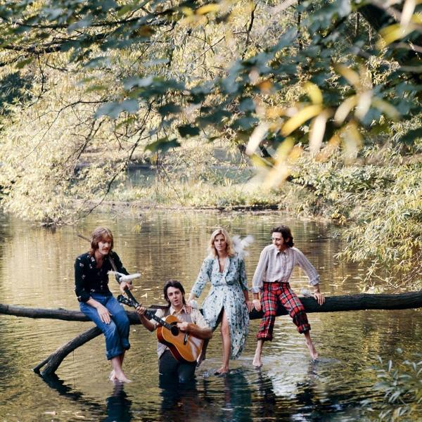 Vier mensen zitten op een boomstam in het water, één speelt gitaar.