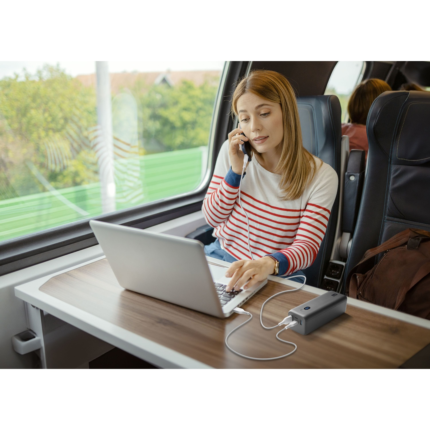 Donna al telefono digita su un laptop in treno. Power bank in carica.