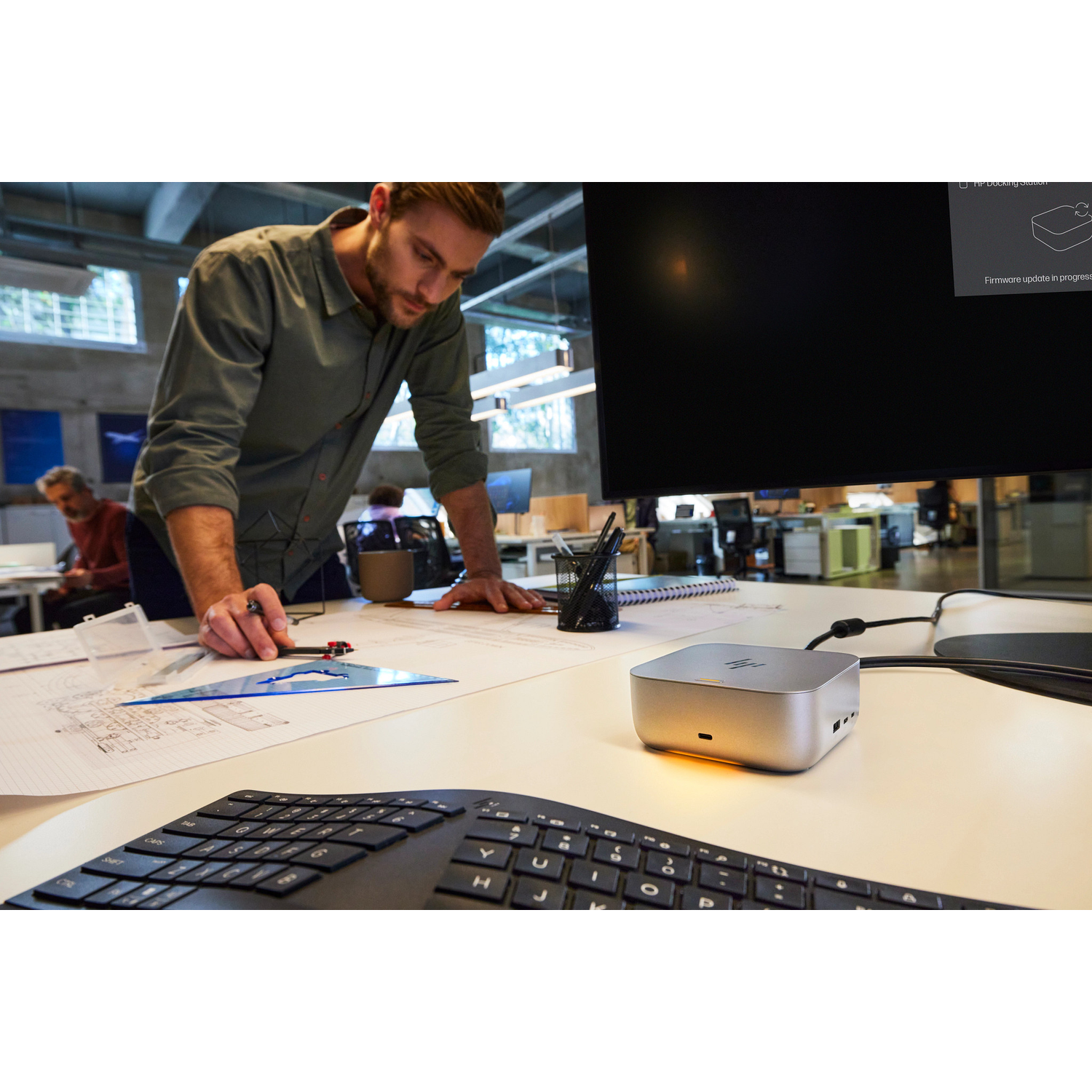 Een man buigt zich over een bureau met blauwdrukken. Een kleine zilveren computer staat ernaast.