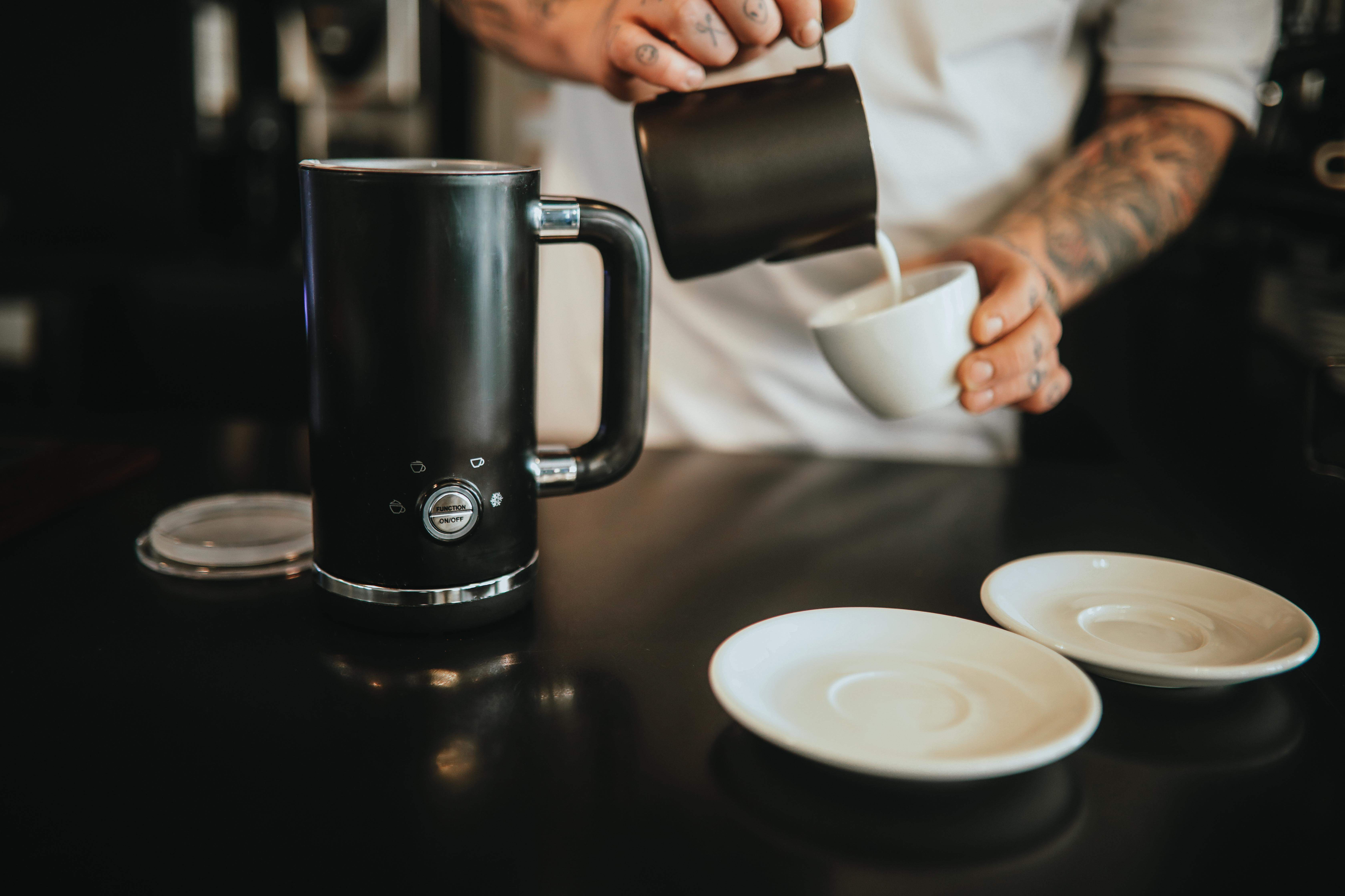 Barista przelewa mleko z czarnego dzbanka do białej filiżanki. Spieniacz mleka obok.