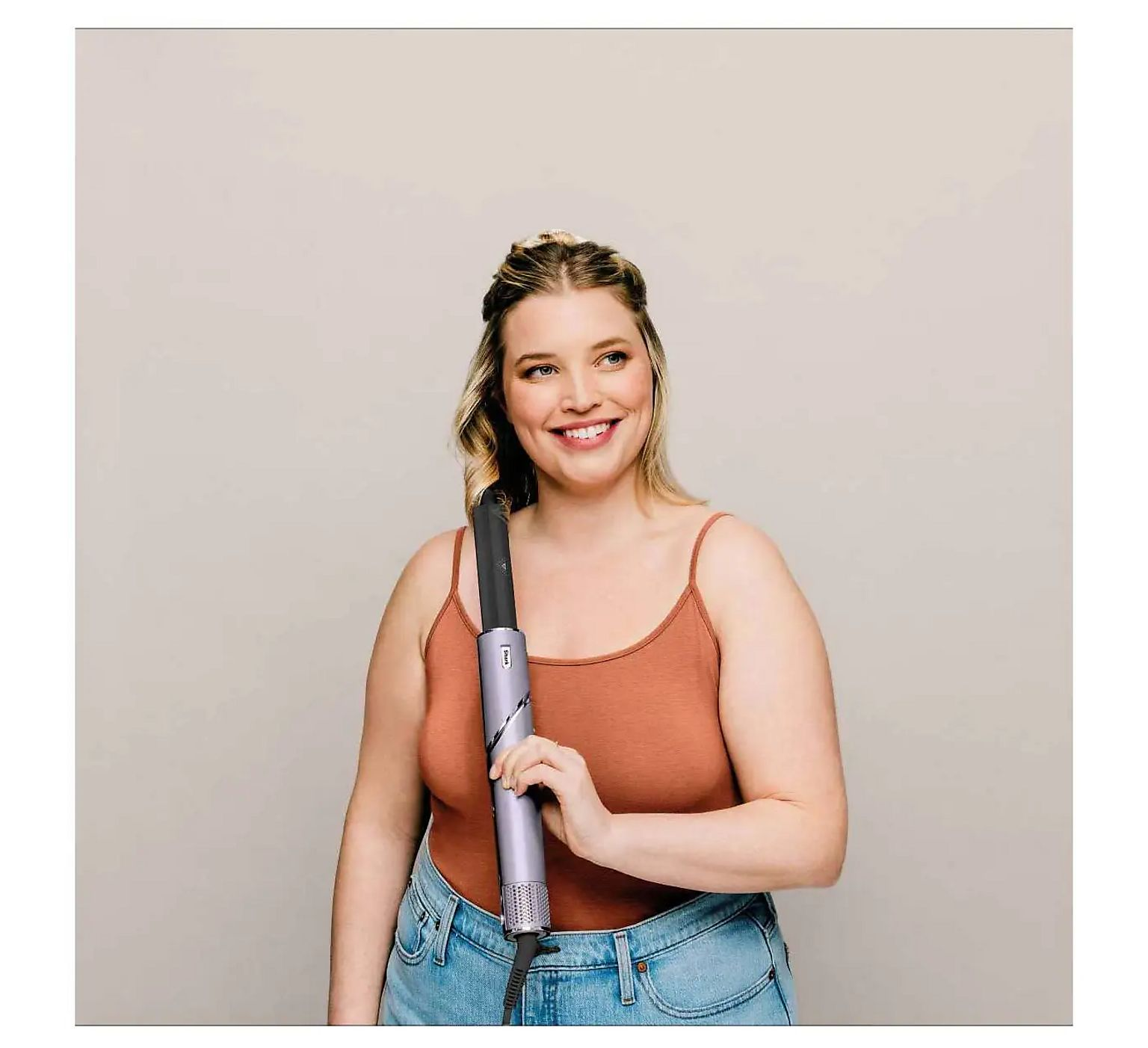 Une femme tient un fer à friser argenté. Elle porte un haut orange et un jean.