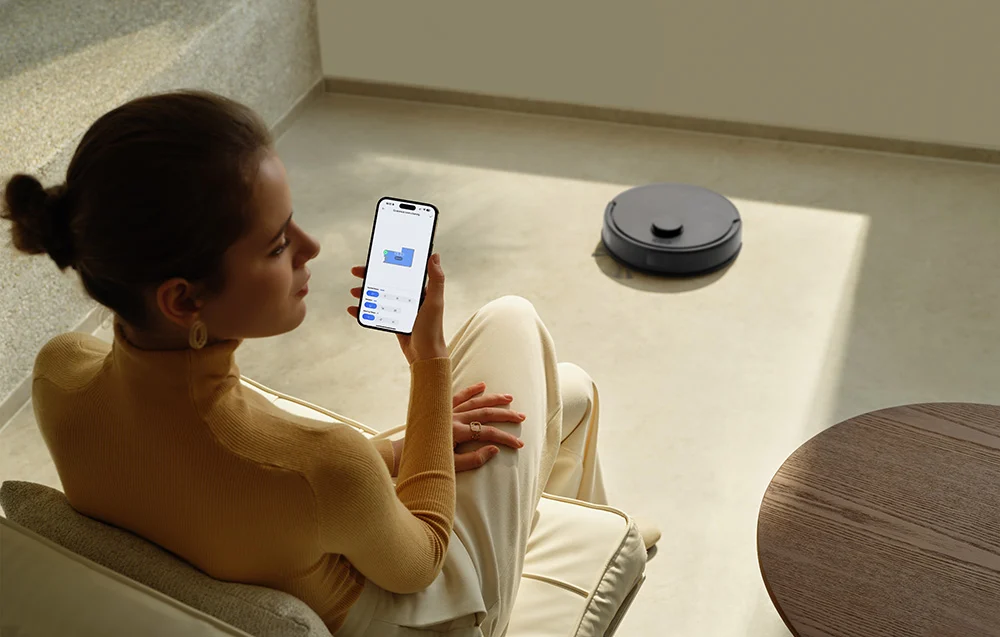 Nő okostelefonnal irányít robotporszívót modern nappaliban.