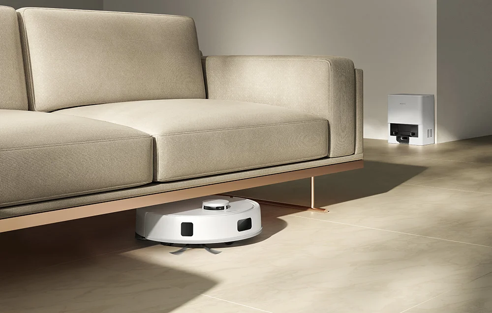 Egy fehér robotporszívó tisztít egy bézs kanapé alatt. A háttérben egy dokkolóállomás látható.