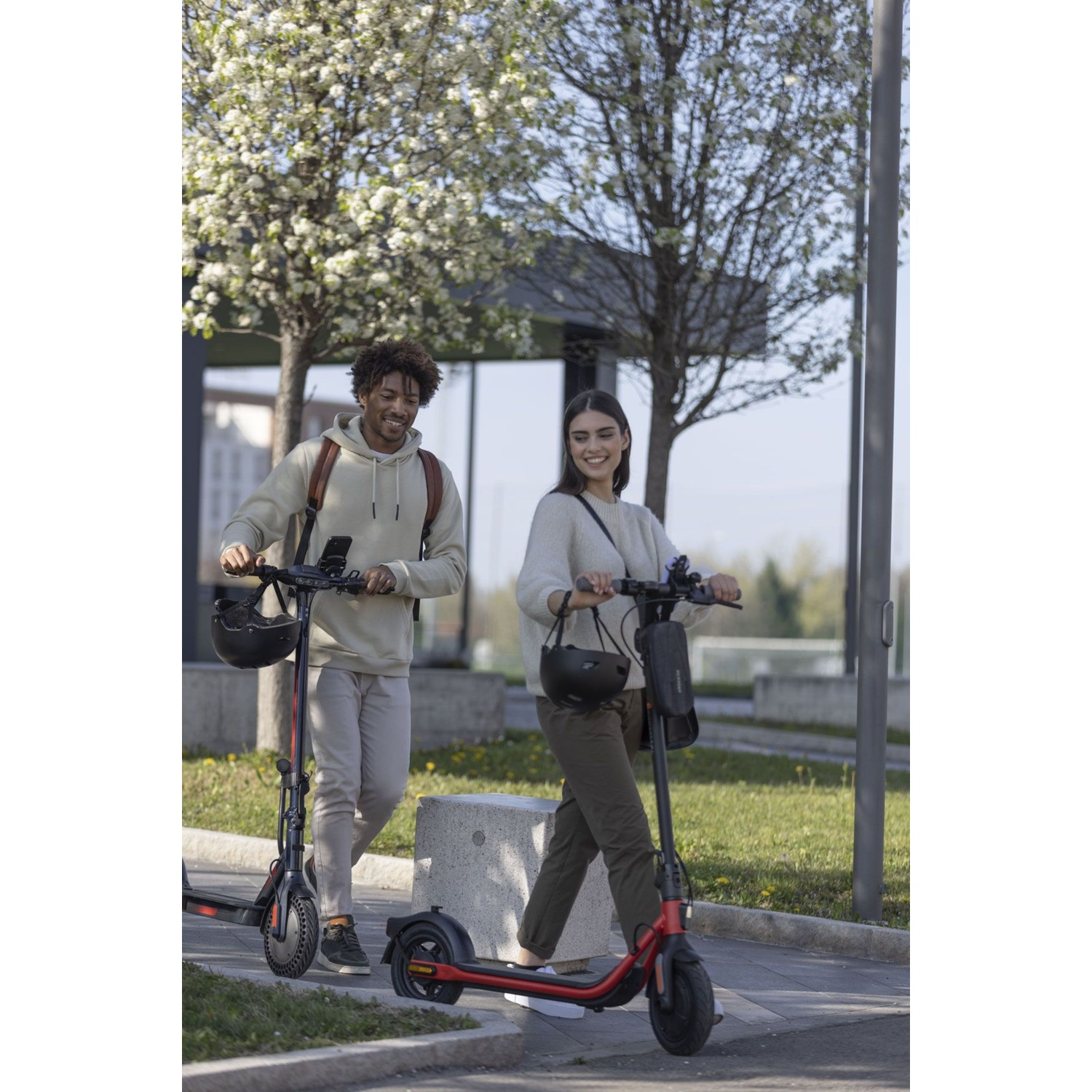 Un uomo e una donna guidano monopattini elettrici su un marciapiede sotto alberi in fiore.