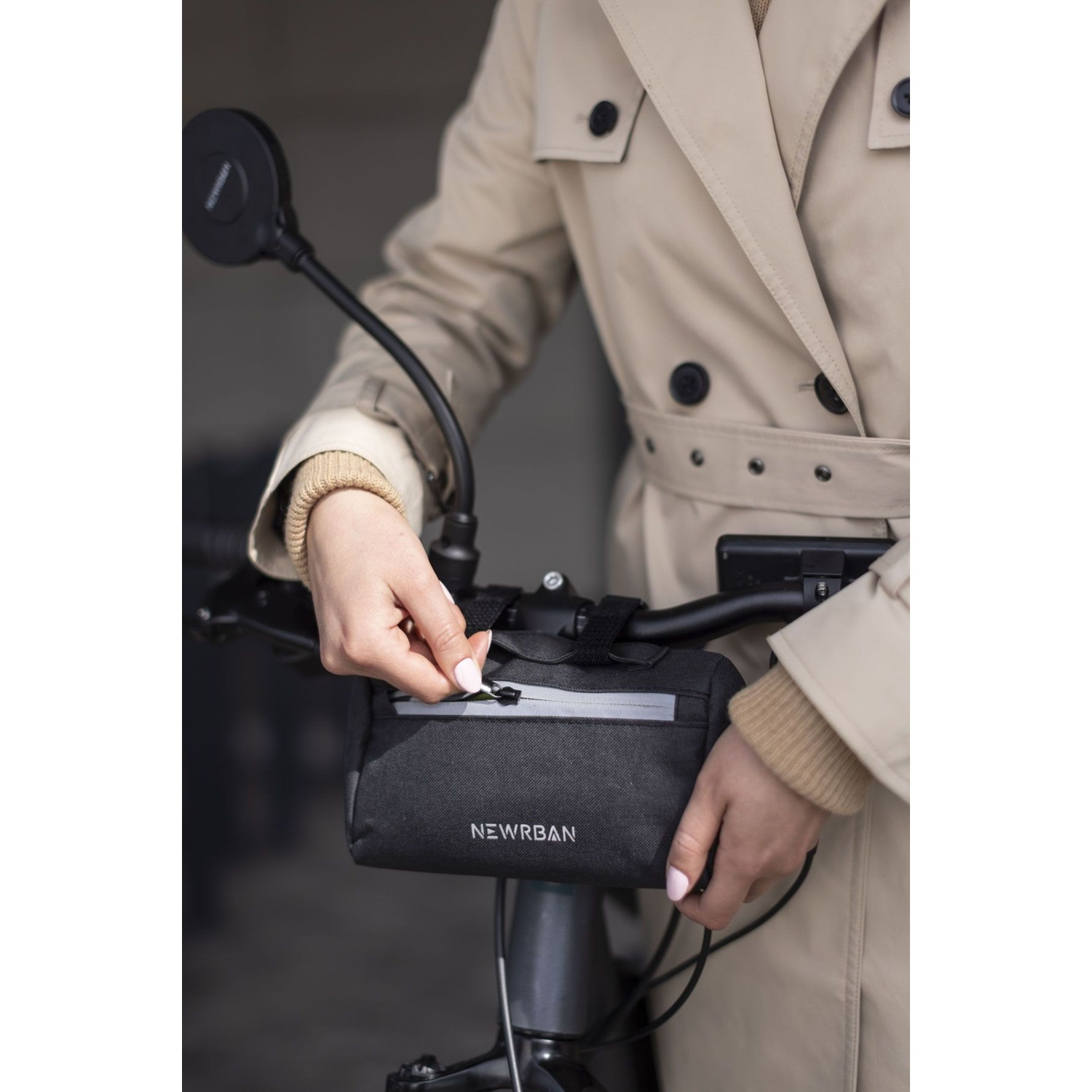 Una persona in trench beige apre una borsa nera sul manubrio di una bicicletta.