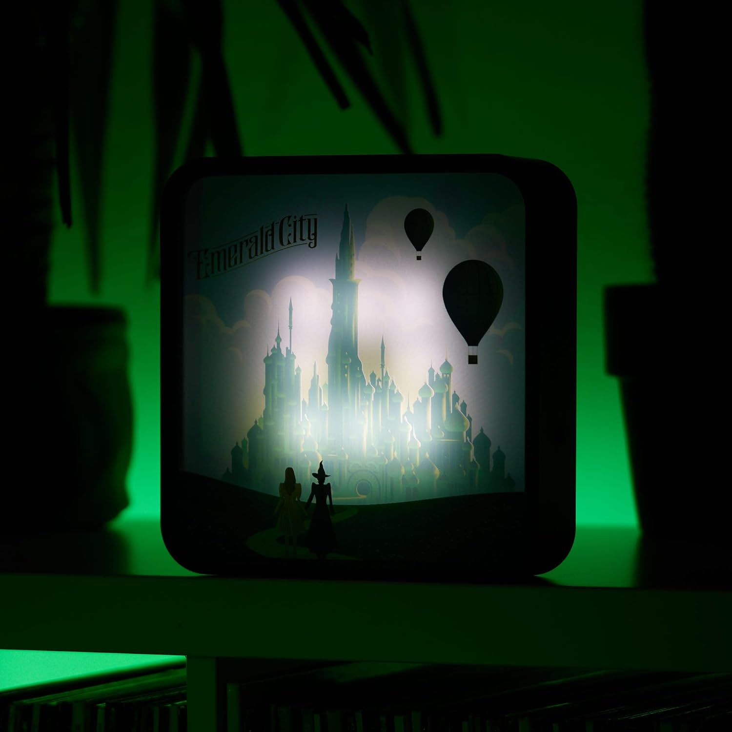 Emerald City két figurával és két léggömbbel az előtérben, városi sziluett a háttérben.