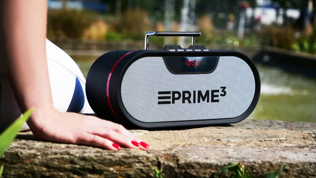 Czarny głośnik z czerwoną ramką i wyświetlaczem, siedzi na kamieniu na zewnątrz.