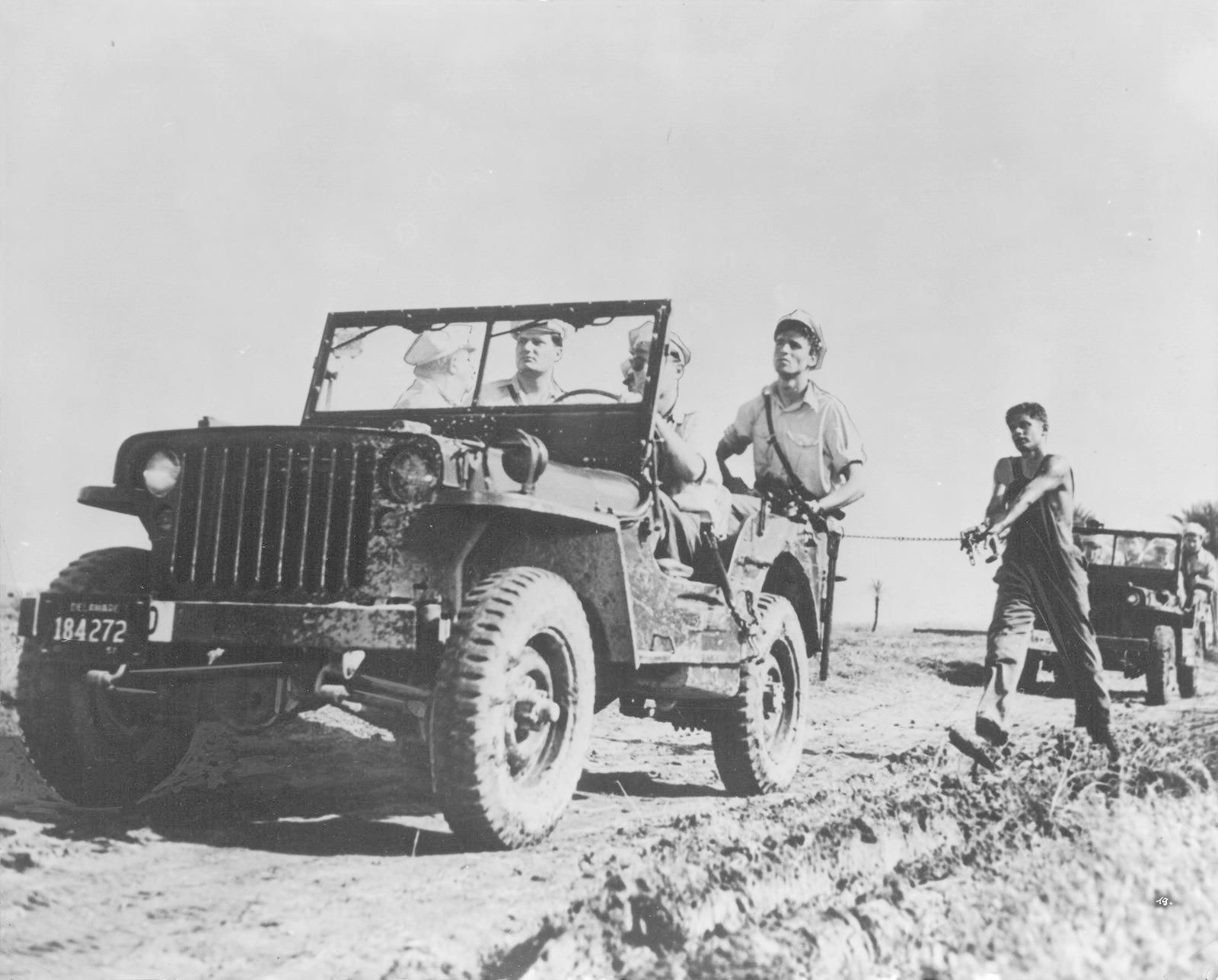 Männer in einem Militärjeep und einem anderen Fahrzeug auf einer unbefestigten Straße. Ein Mann zieht ein Seil.