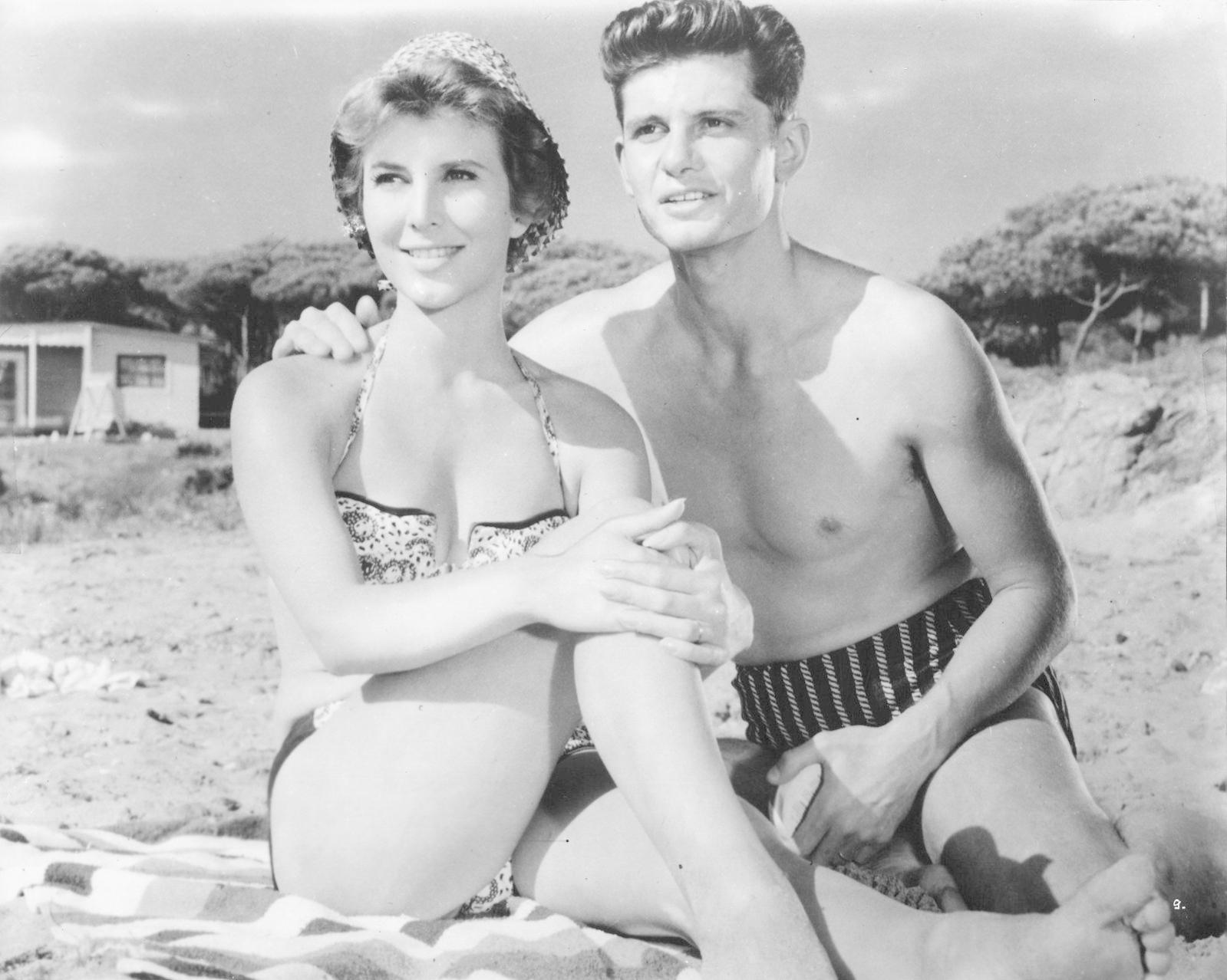Ein Mann und eine Frau in Badekleidung sitzen am Strand vor Bäumen und einem Haus.