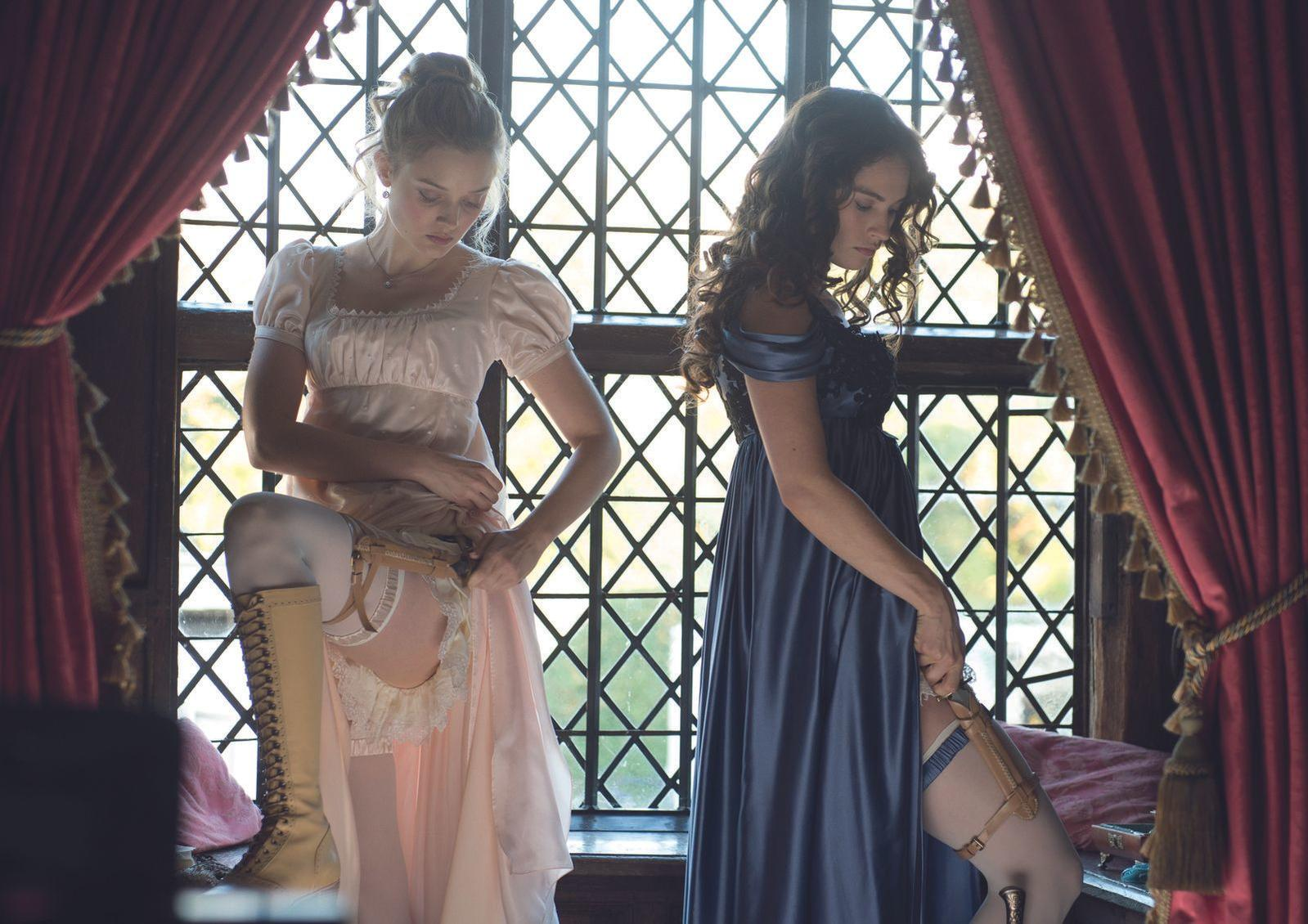 Zwei Frauen in viktorianischer Kleidung stehen am Fenster. Eine trägt ein rosa Kleid, die andere ein blaues.