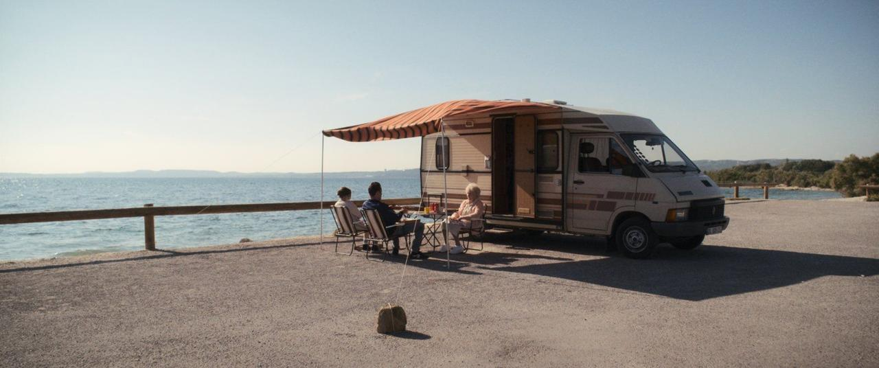 Drei Personen sitzen an einem Tisch vor einem Wohnmobil am Meer.