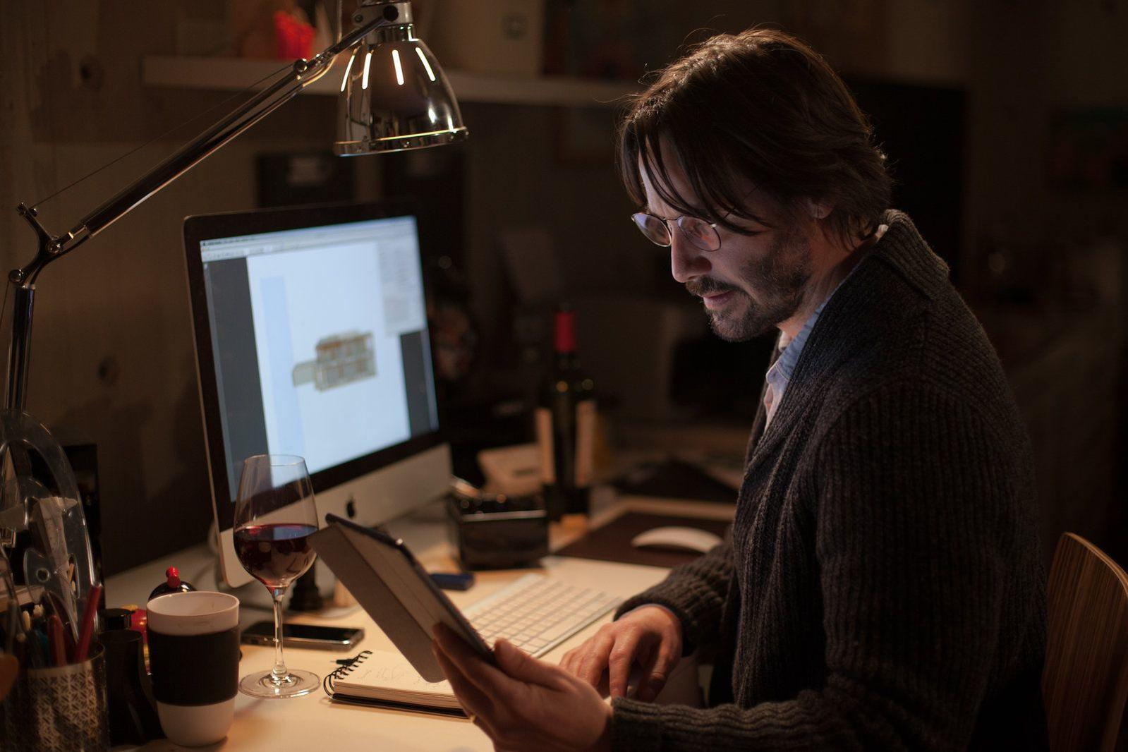 Mann mit Brille arbeitet am Computer, ein Glas Rotwein steht daneben.