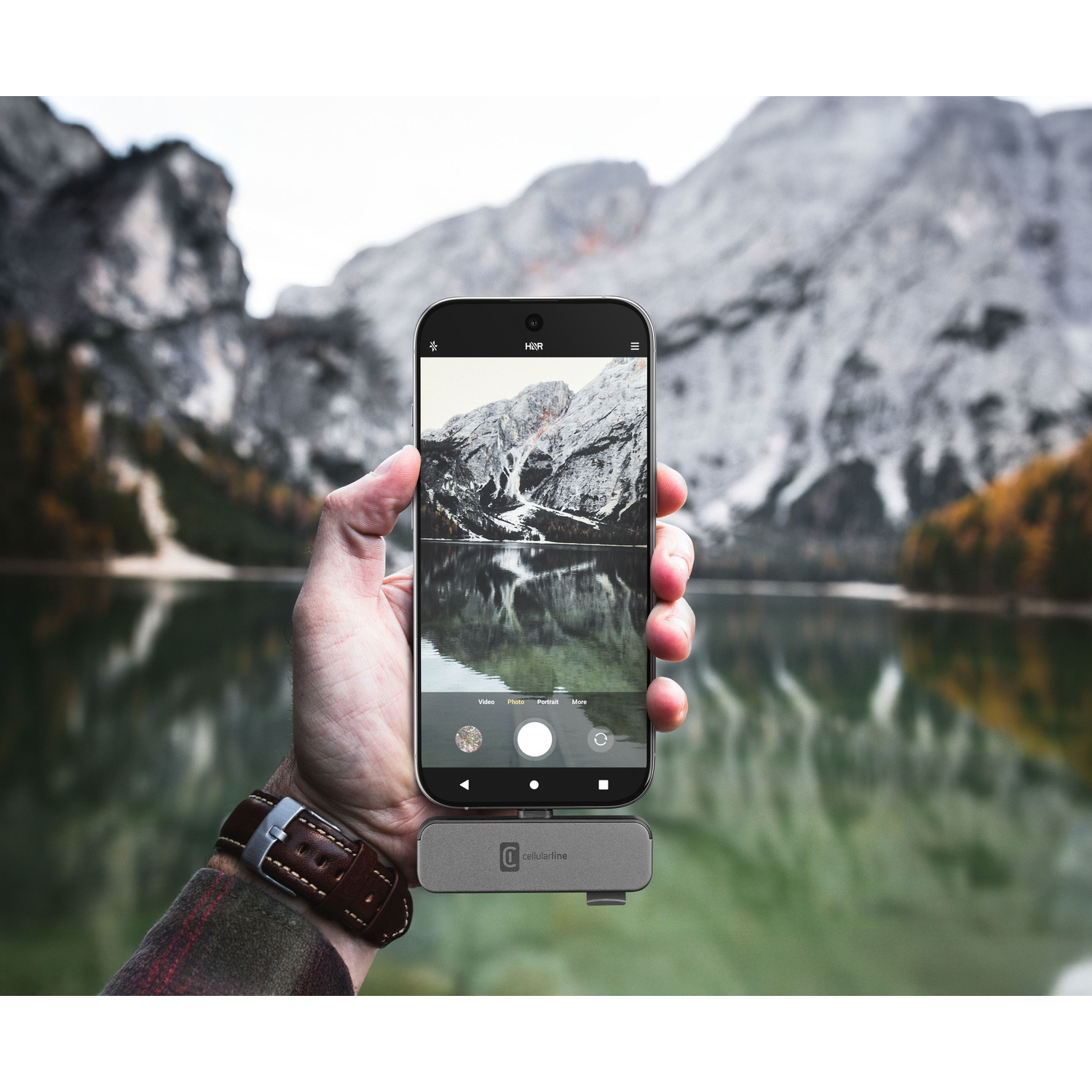 Mano tiene smartphone che fotografa paesaggio montano con lago. Accessorio grigio sotto.