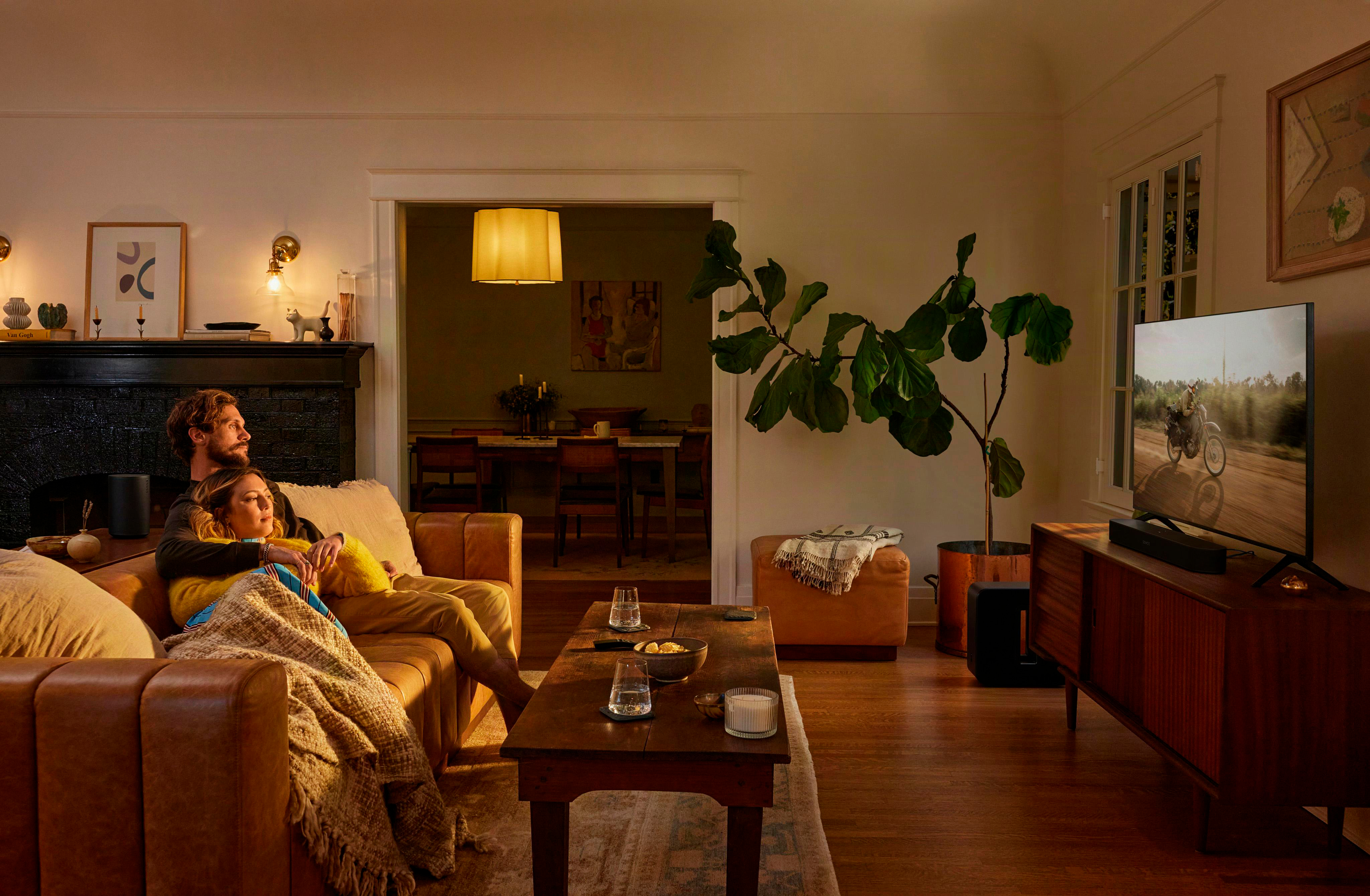 Paar kuschelt auf braunem Sofa, schaut Fernseher. Esszimmer im Hintergrund.
