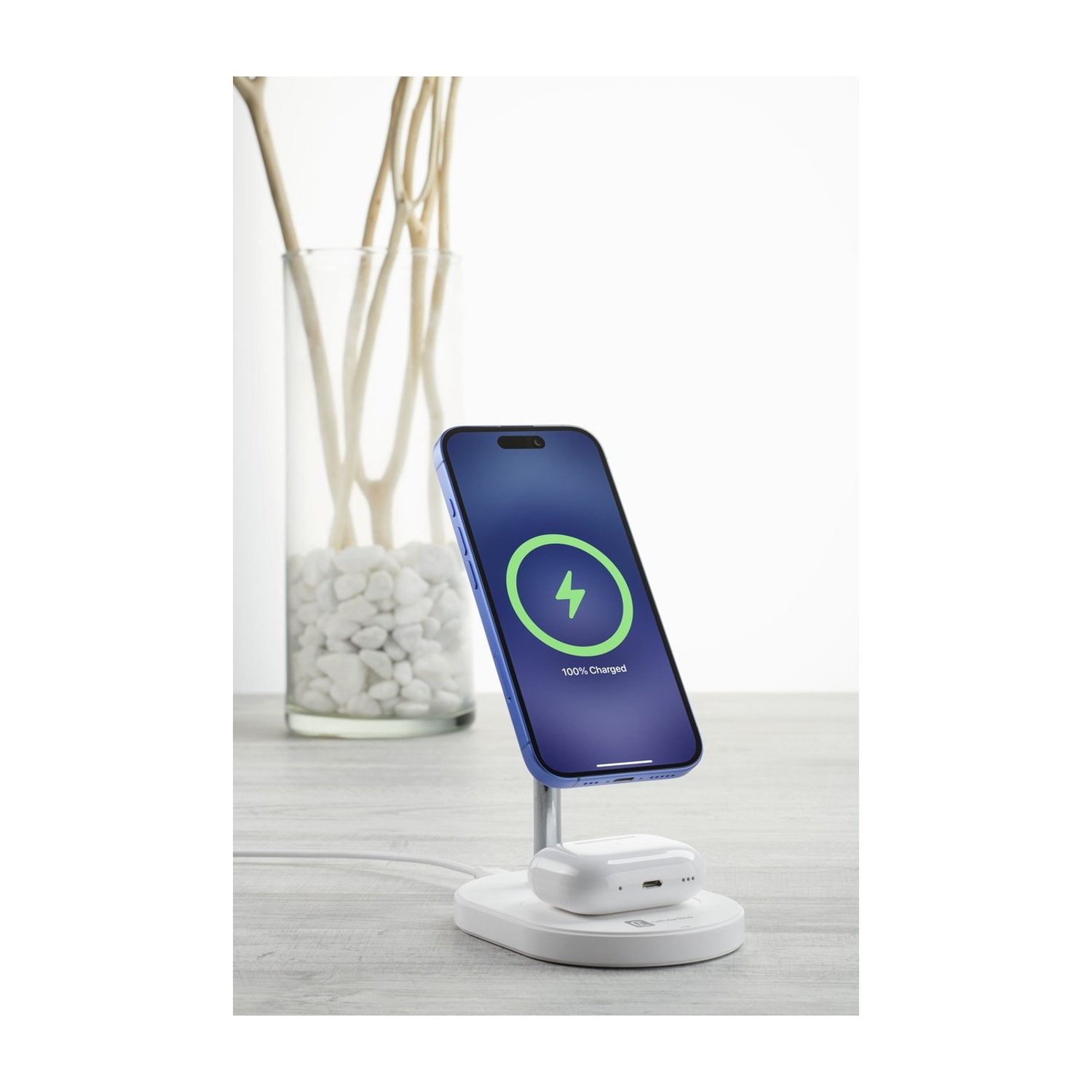 iPhone blu su base di ricarica con AirPods. Sullo sfondo, vaso di vetro con rami.