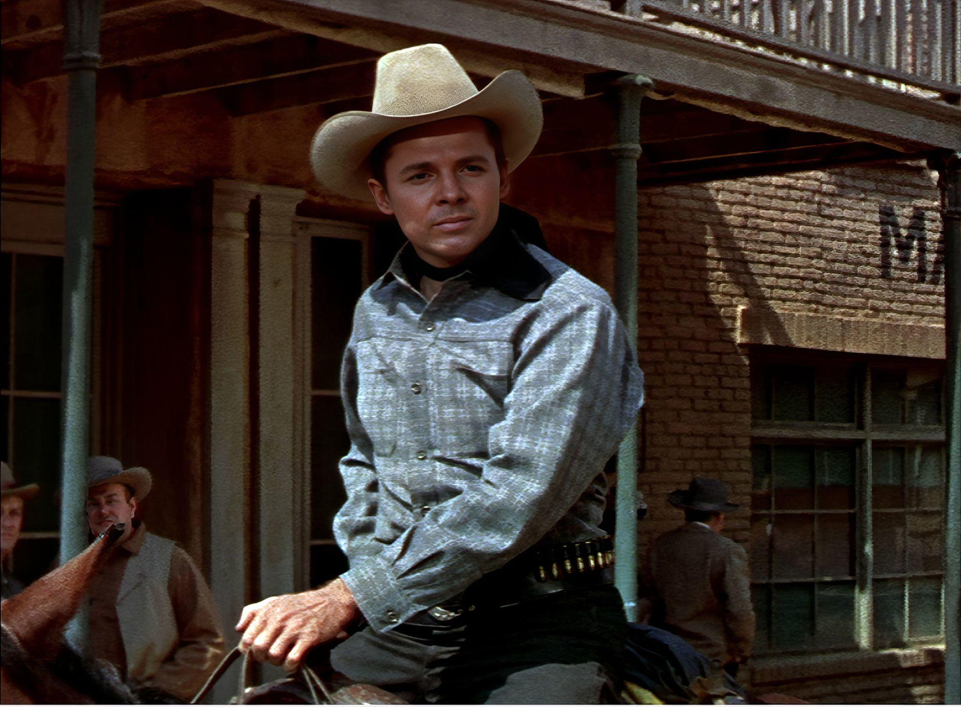 Ein Mann in einem Cowboyhut und kariertem Hemd sitzt auf einem Pferd vor einem Backsteingebäude.