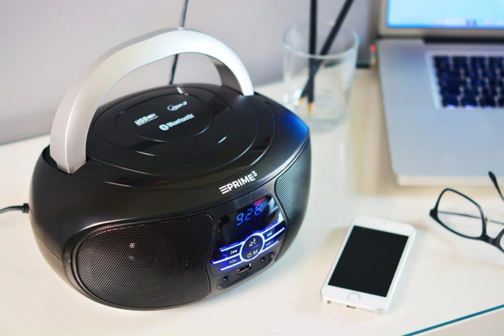 Czarny odtwarzacz CD z srebrnym uchwytem, obok smartfona, laptopa i okularów.