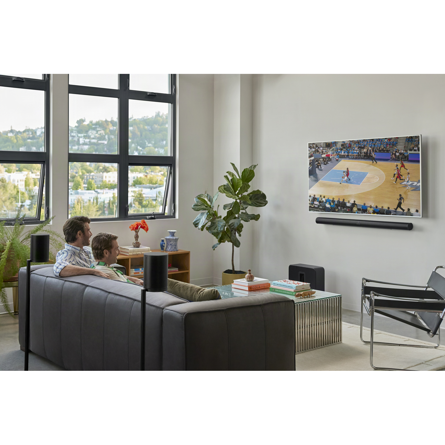 Twee mannen zitten op een bank en kijken basketbal op tv. Een groot scherm toont het spel met spelers.