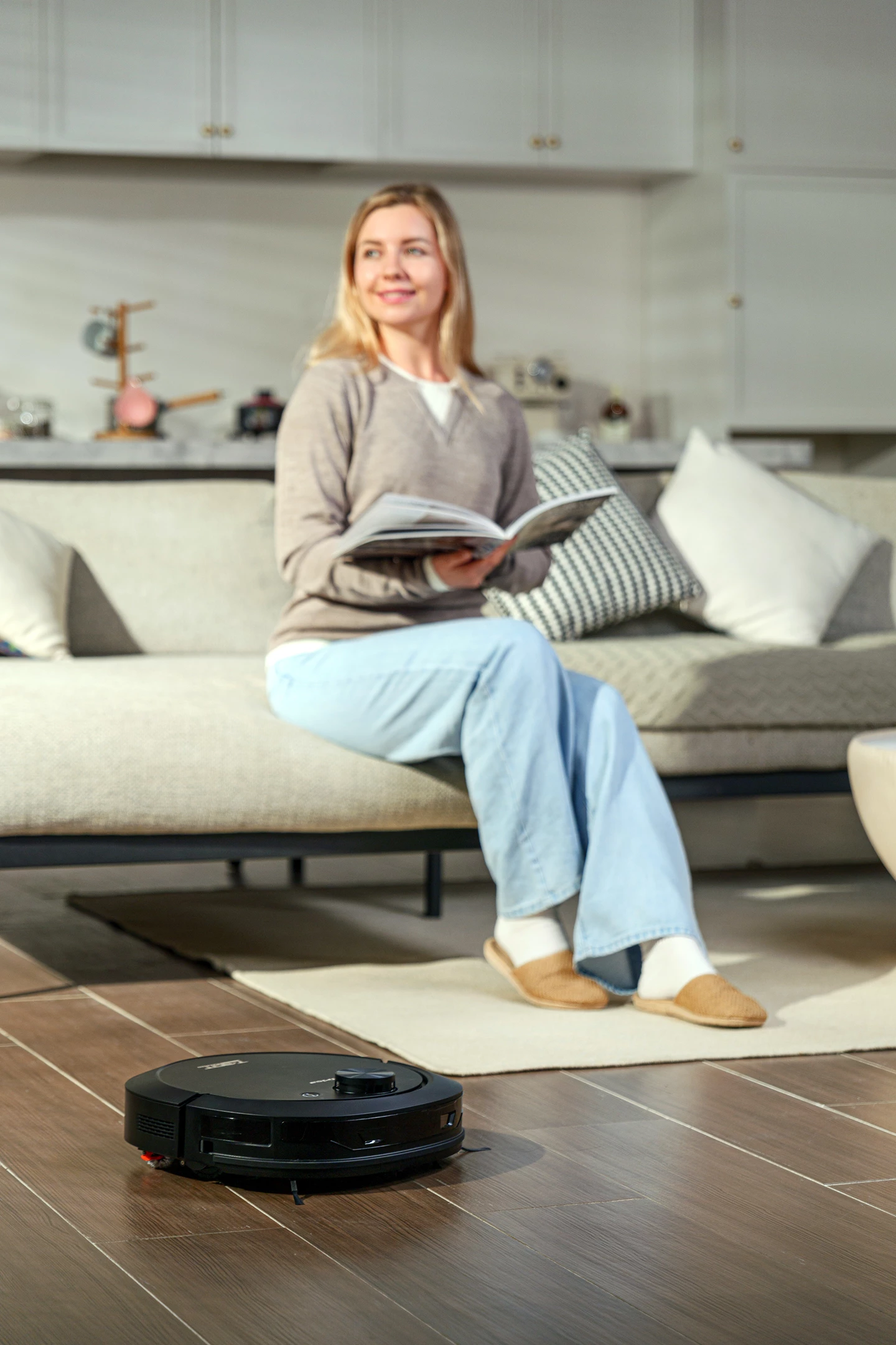 Una donna seduta su un divano legge. Un robot aspirapolvere si muove sul pavimento in legno.