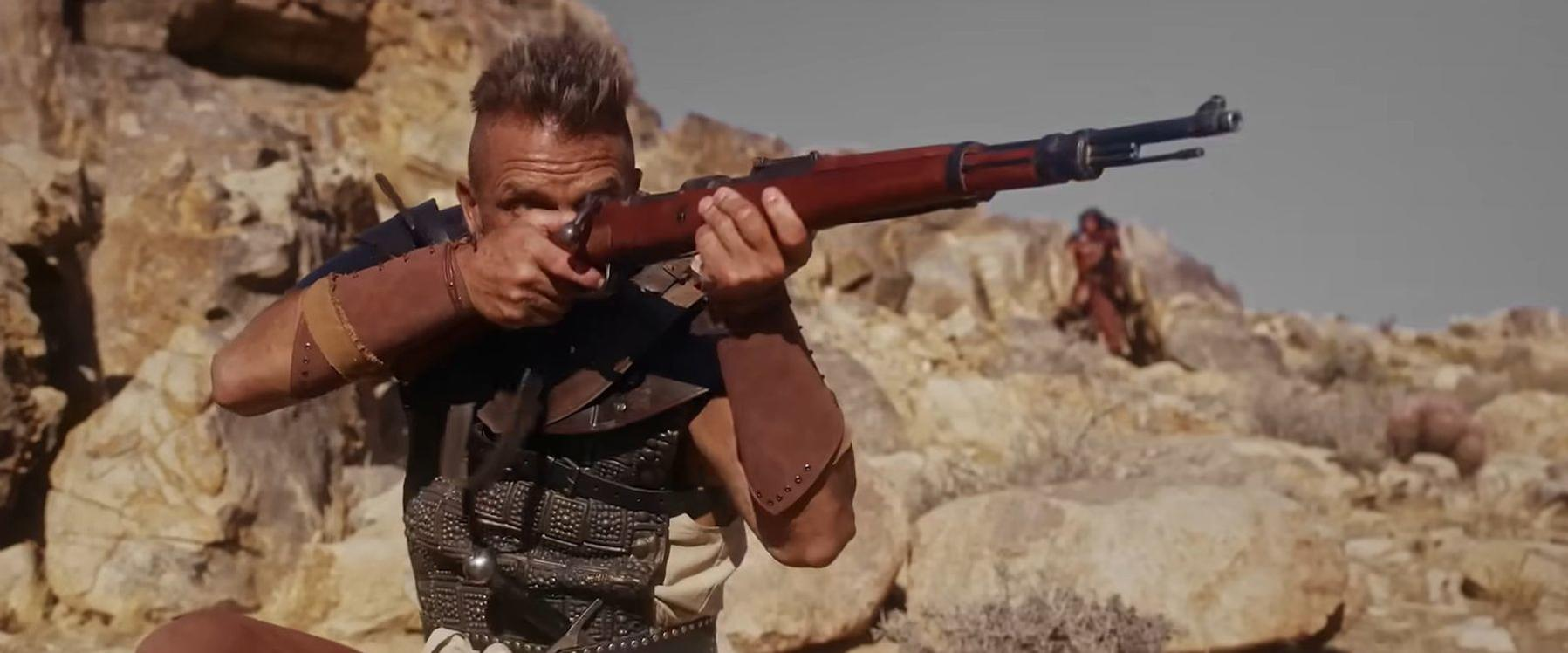 Ein Mann mit Irokesenschnitt zielt mit einem Gewehr in einer felsigen Wüstenlandschaft.