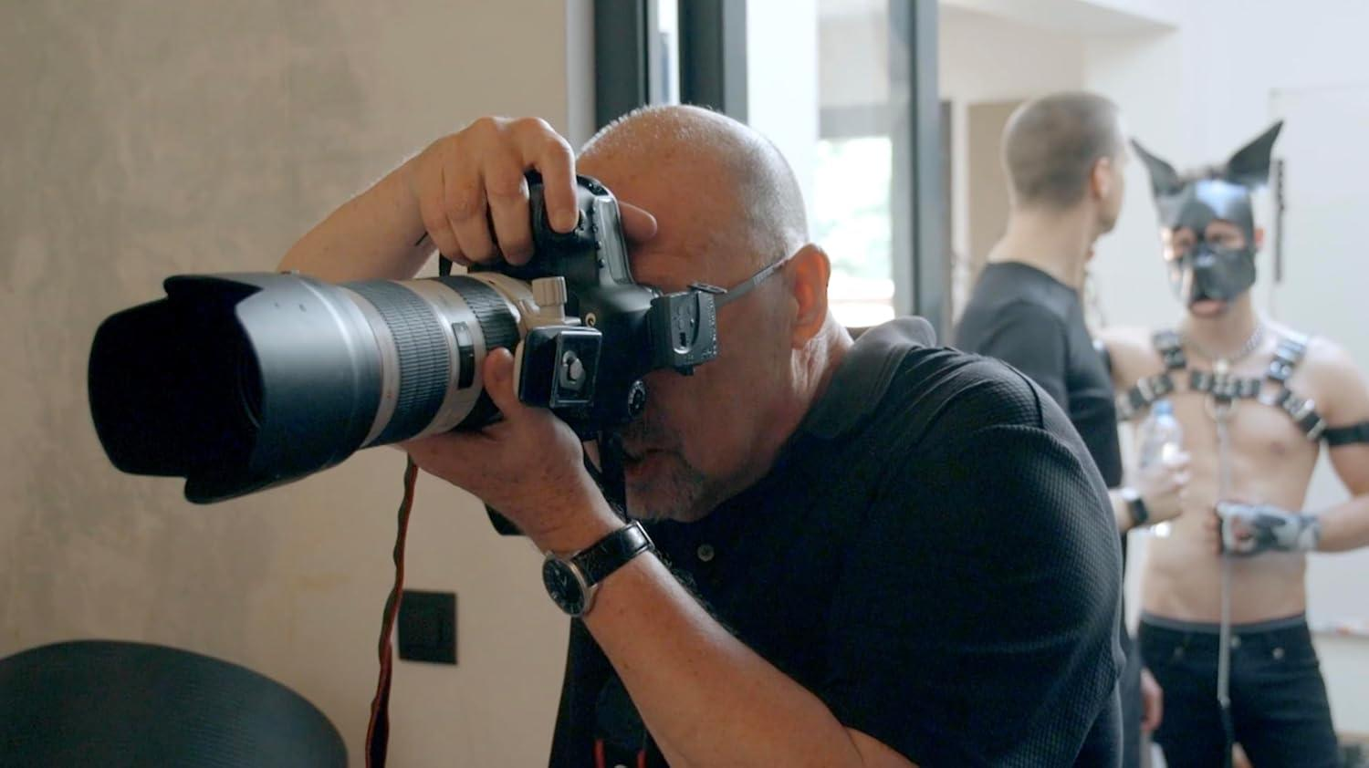 Ein Mann mit Kamera fotografiert, im Hintergrund eine Person mit Maske.