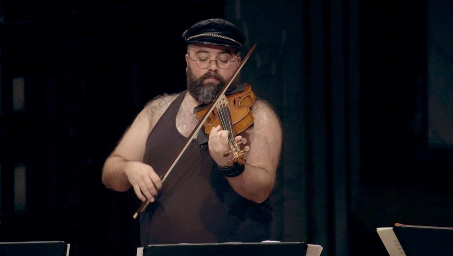 Ein Mann mit Bart und Brille spielt Geige, trägt eine schwarze Kappe und ein Tanktop.