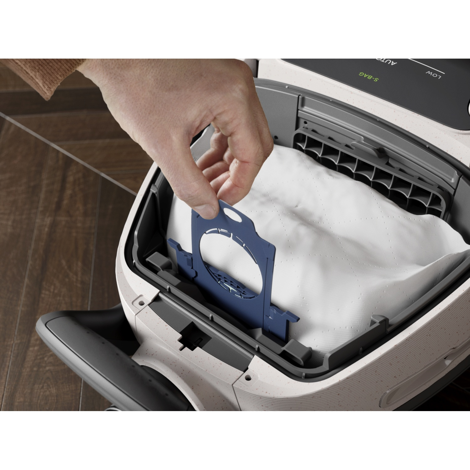 A hand inserts a blue holder into a vacuum cleaner bag.