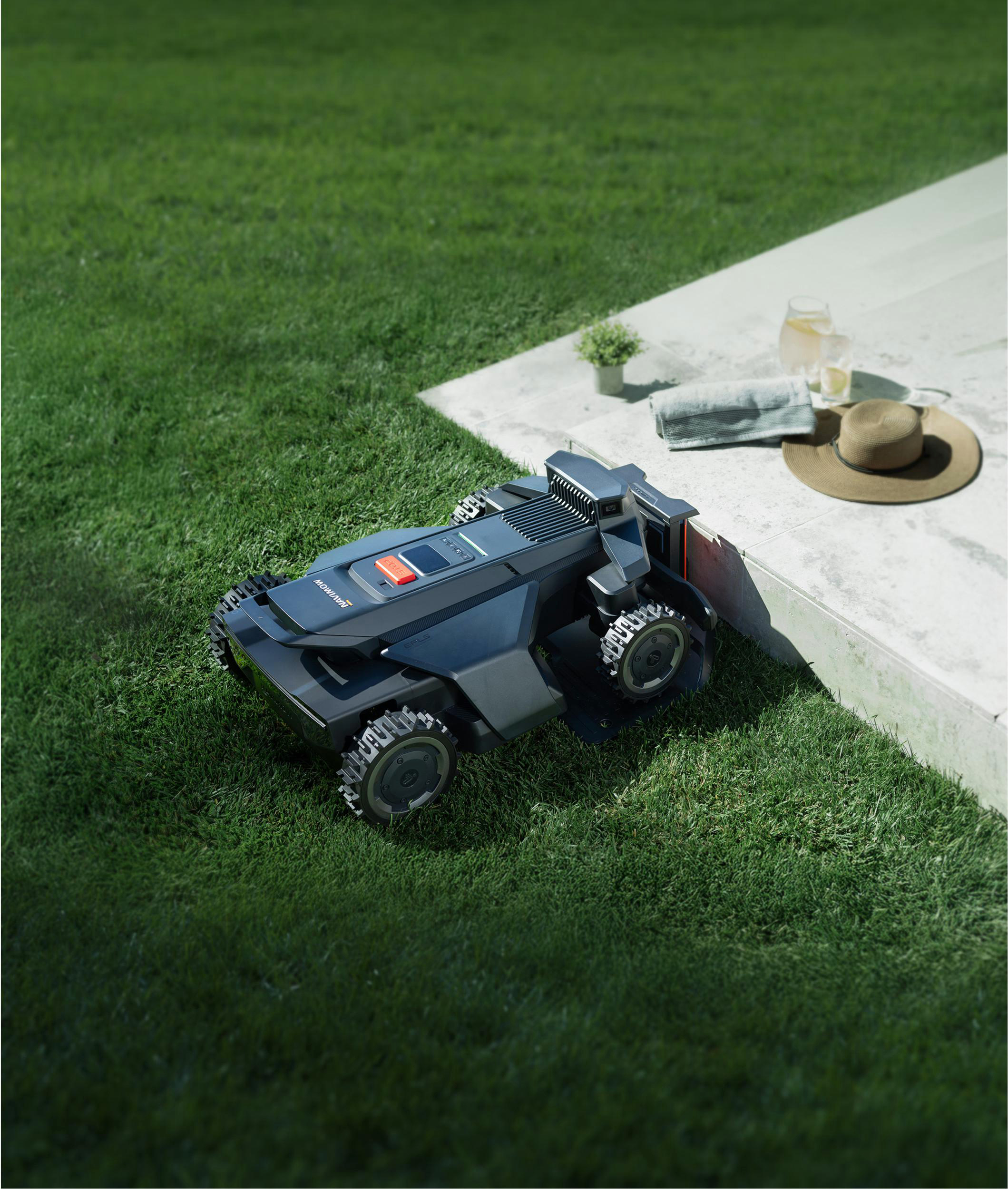 Ein Mähroboter steht auf einem Rasen neben einer Betonfläche mit Hut und Getränken.