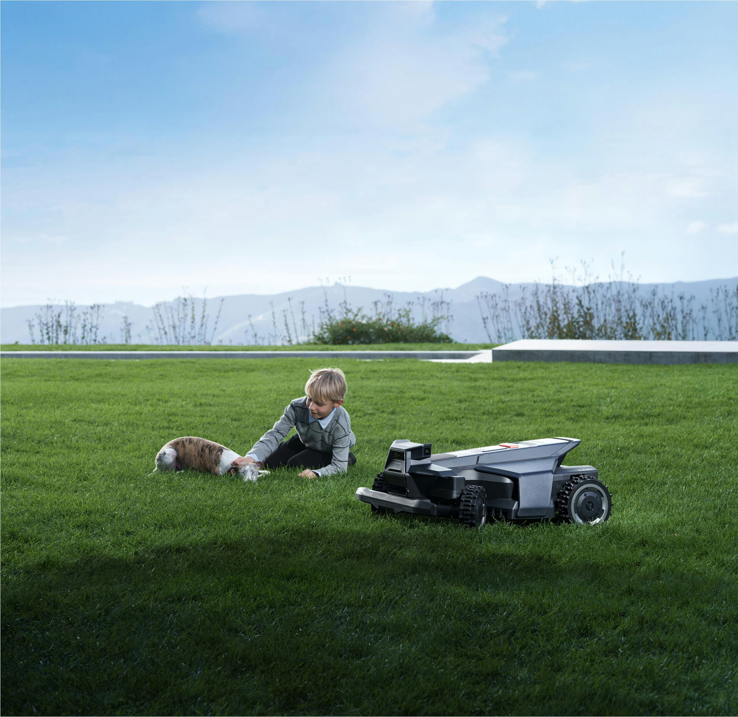 Ein Junge spielt mit einem Hund auf dem Rasen neben einem Mähroboter.