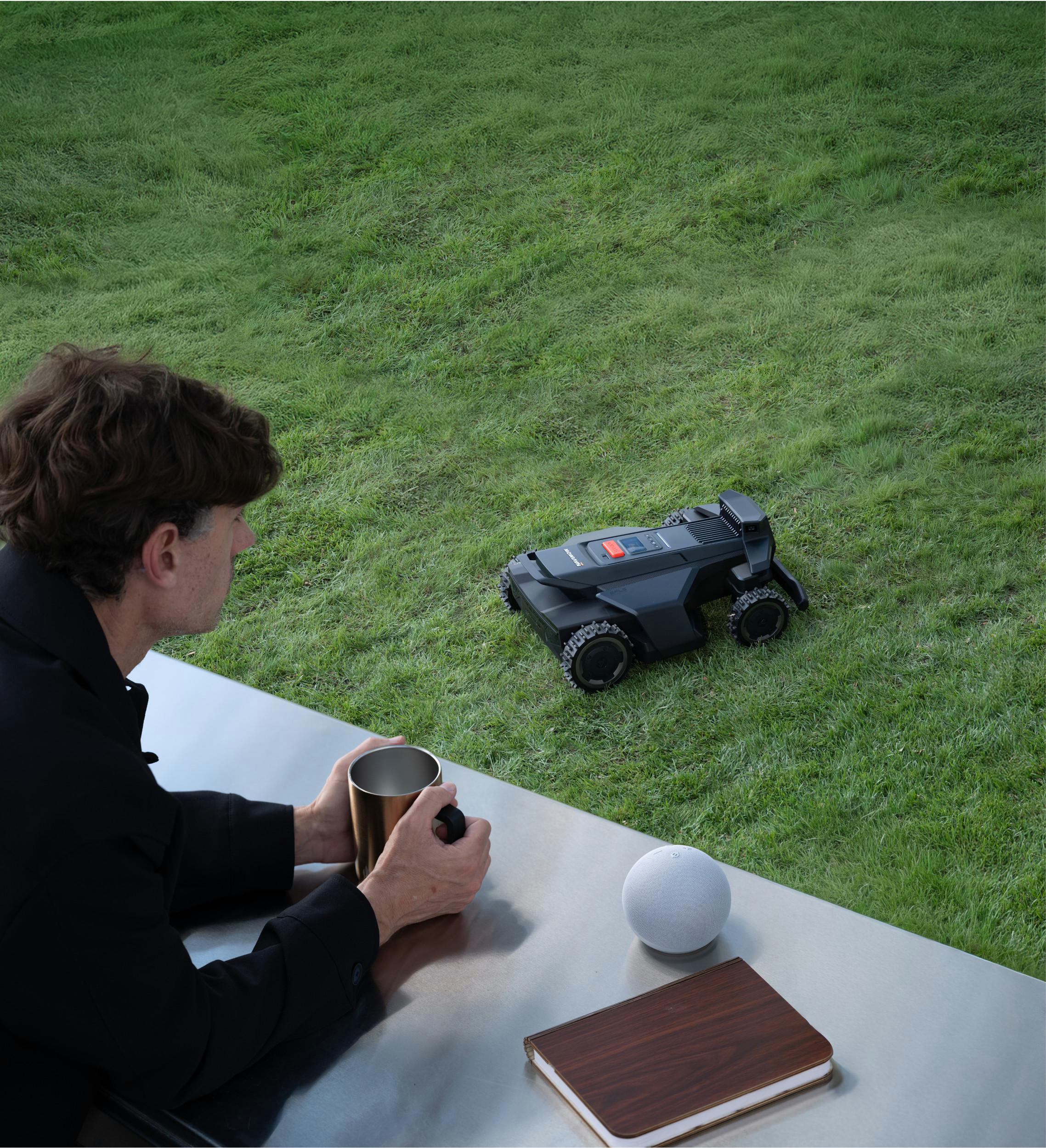 Mann hält Tasse, Rasenmäherroboter und Notizbuch auf Tisch im Gras.
